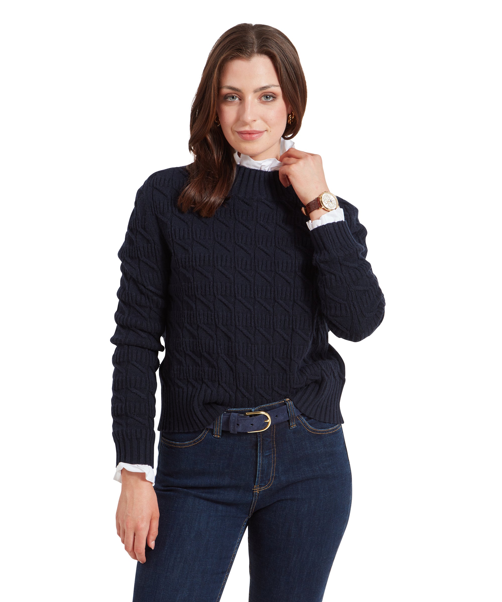 The woman is wearing a Schöffel Sandsend Cable Jumper for Women in Navy paired with dark blue jeans. She has a white ruffled shirt underneath the sweater, and her hand is gently resting near her chin. She is smiling softly.