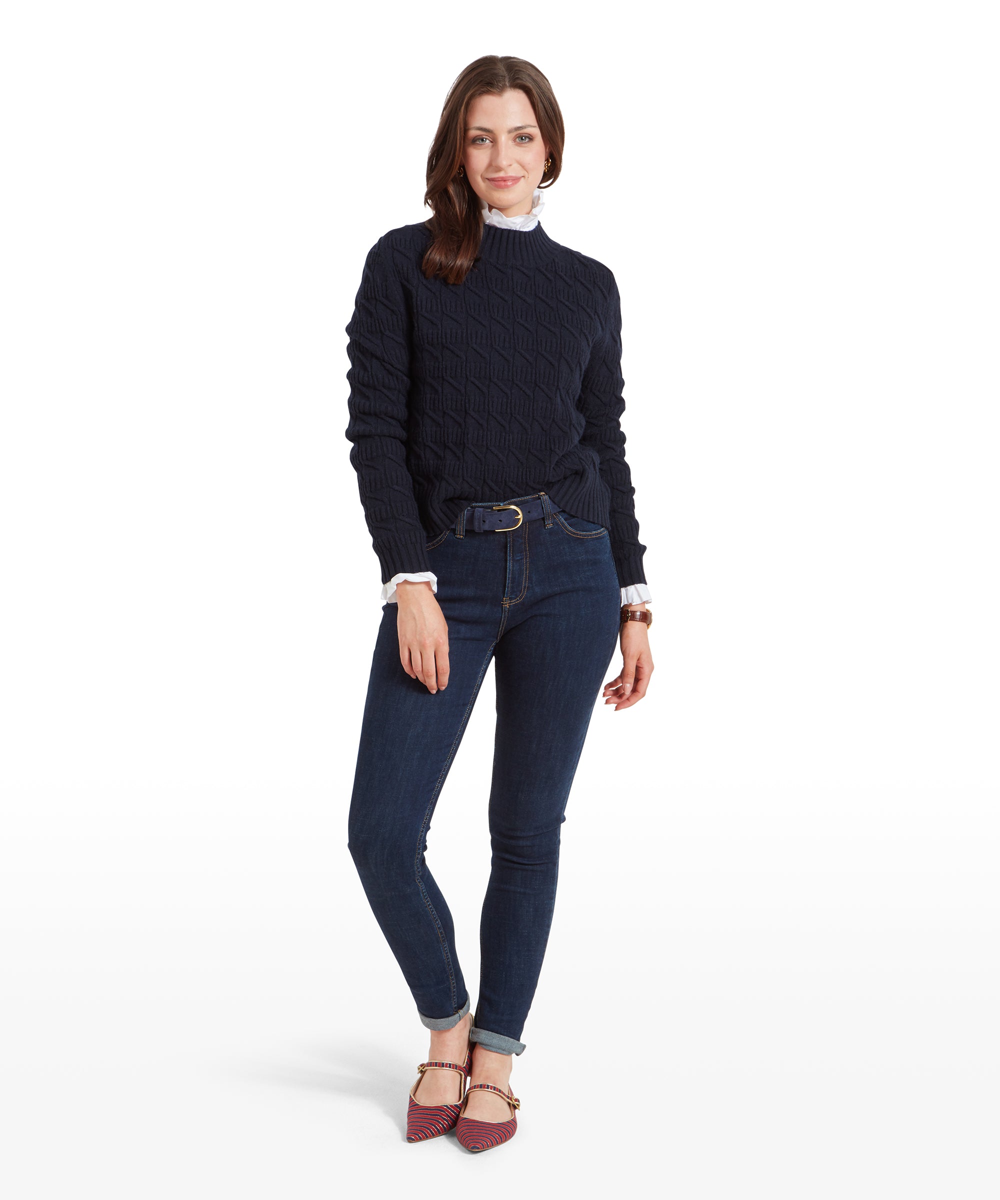 The woman is wearing a Schöffel Sandsend Cable Jumper for Women in Navy with dark blue jeans and red striped shoes. She is standing casually with one hand in her pocket and the other resting on her thigh, smiling gently.