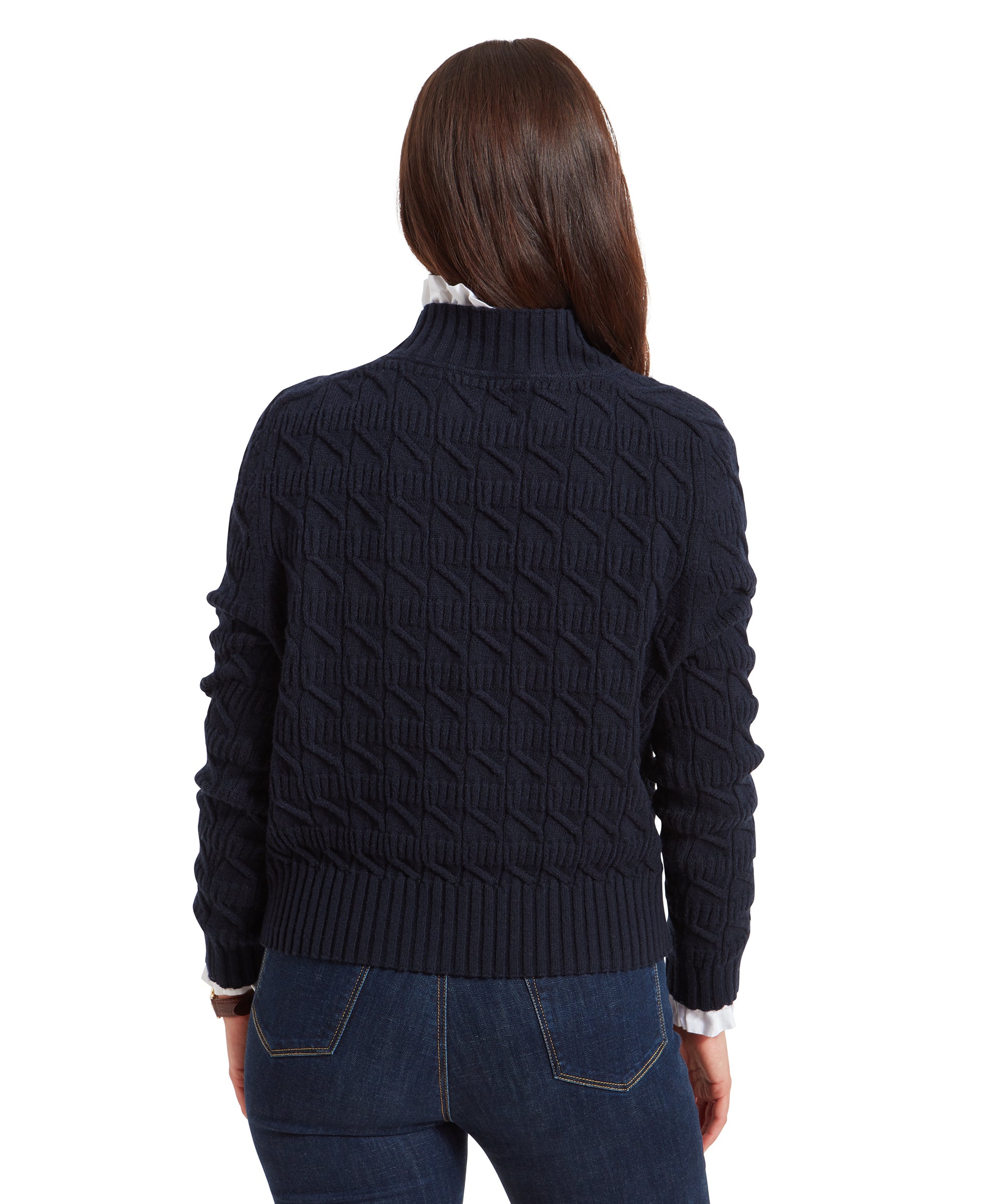 The back of the Schöffel Sandsend Cable Jumper for Women in Navy is visible, showing the intricate pattern. The woman is also wearing dark blue jeans, and her hair is styled neatly.