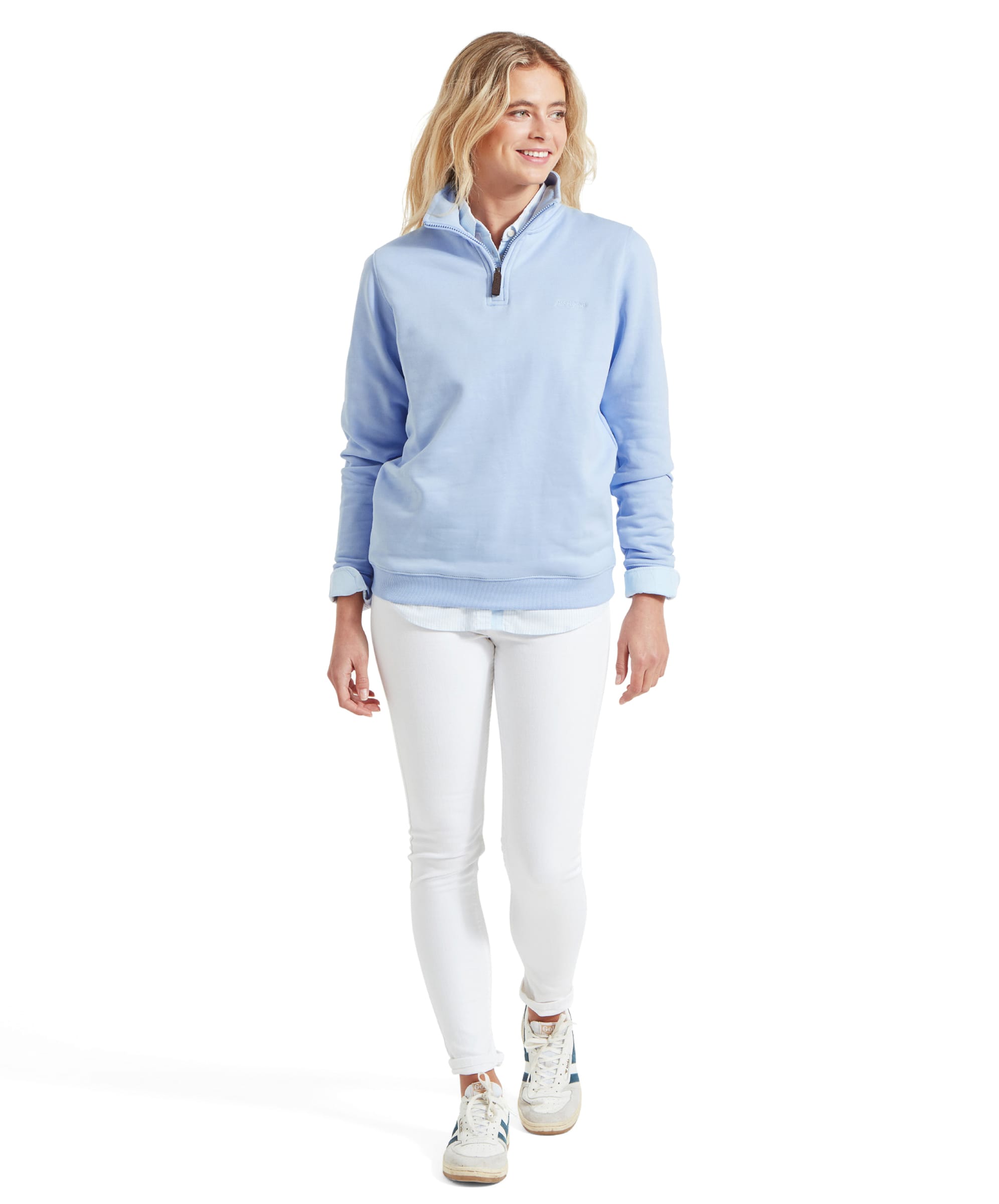 The same woman is shown standing and walking forward, dressed in a Schöffel Sennen Cove Sweatshirt for Womens in Blue, white jeans, and white trainers with blue details.