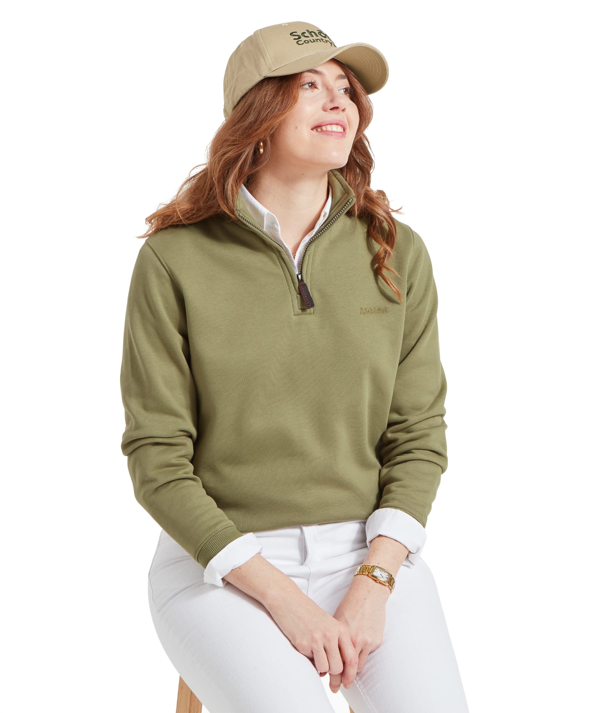 Woman sitting and smiling, wearing a Schöffel Sennen Cove Sweatshirt for Womens in Green with a beige cap, paired with white jeans.