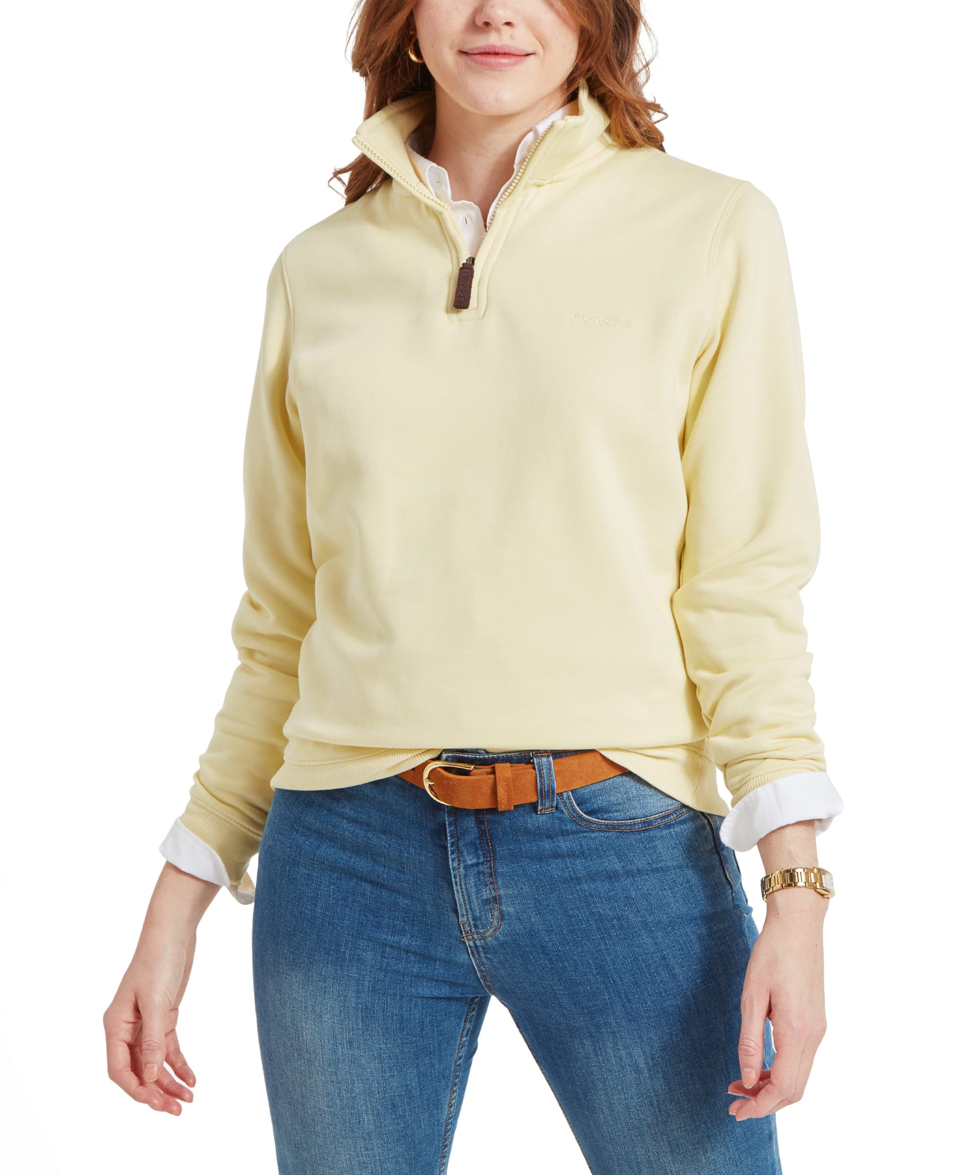 A woman wearing a Schöffel Sennen Cove Sweatshirt for Womens in Yellow with a white collared shirt underneath. She is standing with one hand slightly tucked in her jeans pocket, showcasing the front of the sweatshirt. The outfit is paired with blue jeans and a brown belt, complementing her casual yet polished look.