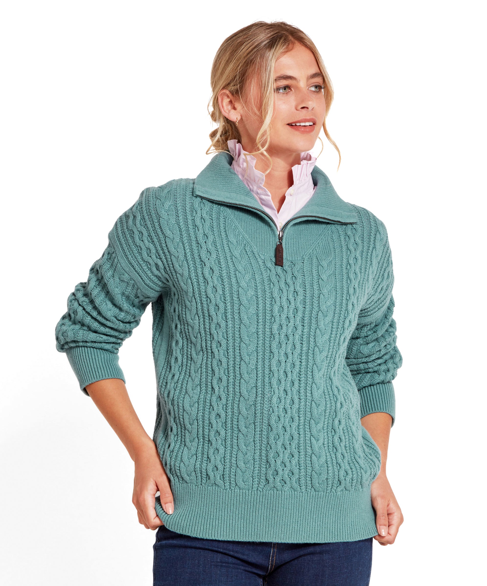 The model is wearing a Schöffel Settle 1/4 Zip for Women in Blue. The sweater features intricate cable patterns and a ribbed hem and cuffs. She pairs it with a light pink ruffled blouse underneath, adding a touch of femininity. Her hands rest in the pockets of her dark blue jeans.