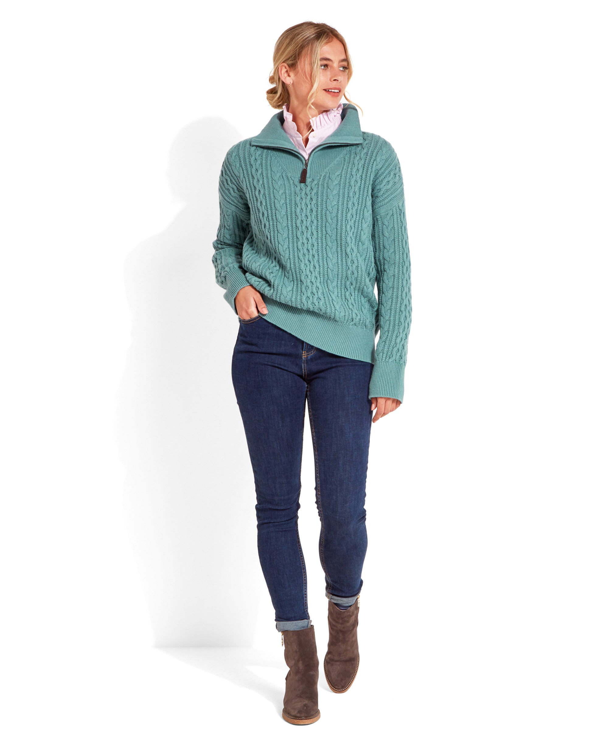 The model is dressed in the Schöffel Settle 1/4 Zip for Women in Blue and dark blue jeans. She completes the look with brown ankle boots. Her hair is styled in loose waves, and she walks confidently with one hand in her pocket.