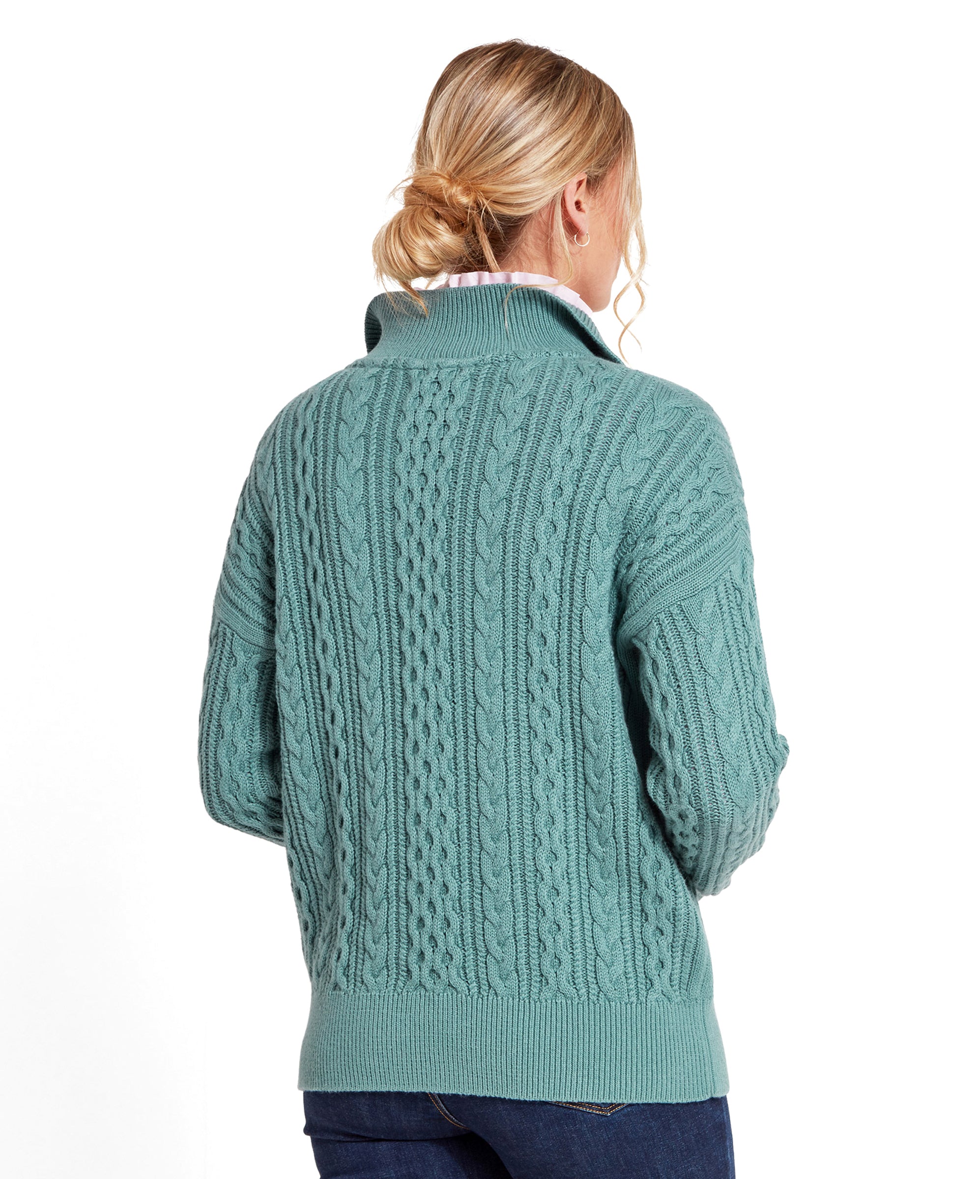 The image shows the back of the Schöffel Settle 1/4 Zip for Women in Blue, highlighting the detailed cable patterns. The model's hair is pulled back in a loose bun, giving a clear view of the sweater's design. She pairs it with dark blue jeans.