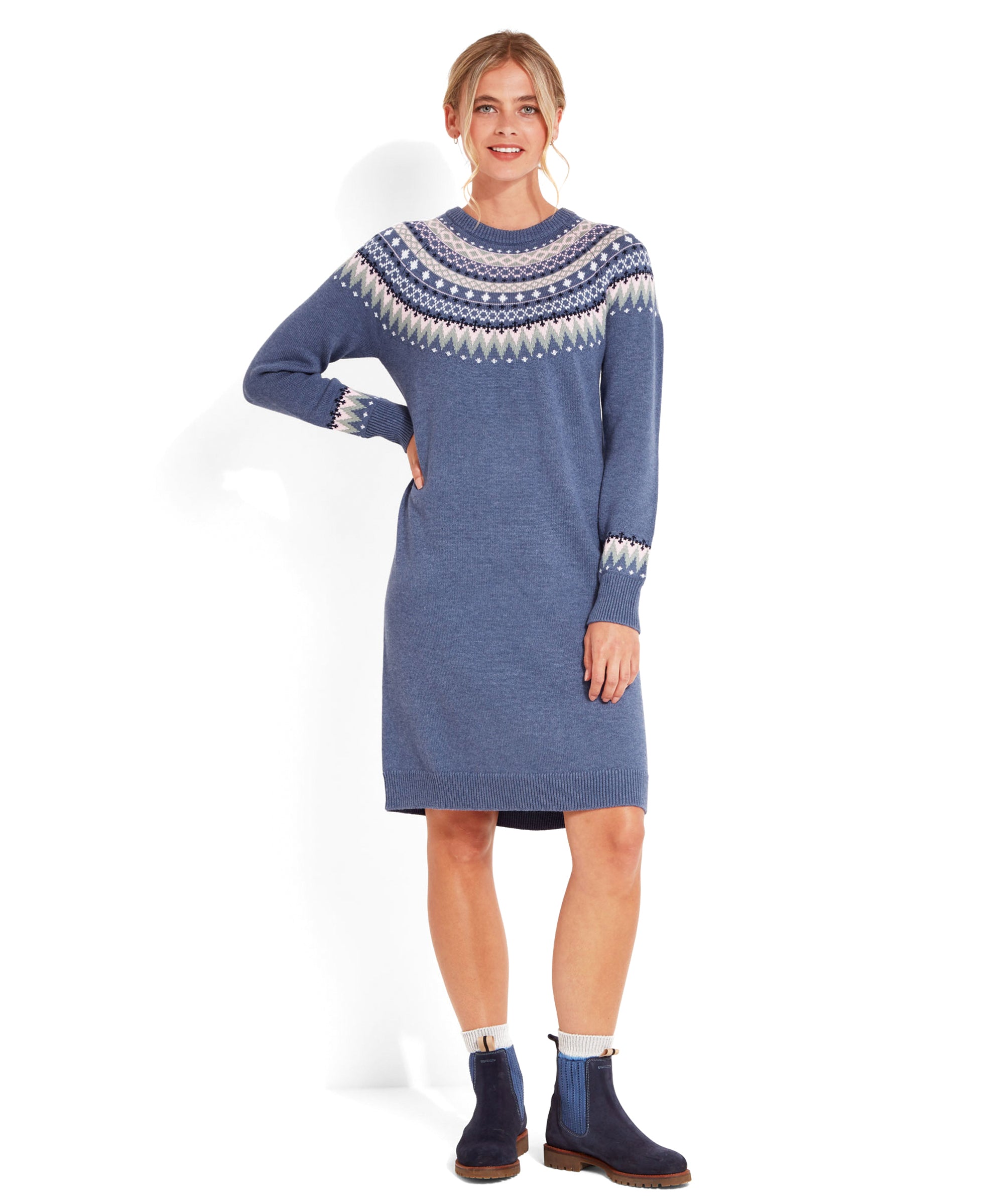 A woman is wearing a Schöffel Shetland Fairisle Dress for Women in Blue with a detailed Fair Isle pattern across the yoke, chest, and sleeves. The pattern includes shades of pink, white, and green, forming intricate geometric shapes. The dress has long sleeves and reaches to her knees. She pairs the dress with blue ankle boots and white socks peeking out from the boots. Her hair is styled in a loose, low bun, and she is standing and smiling.