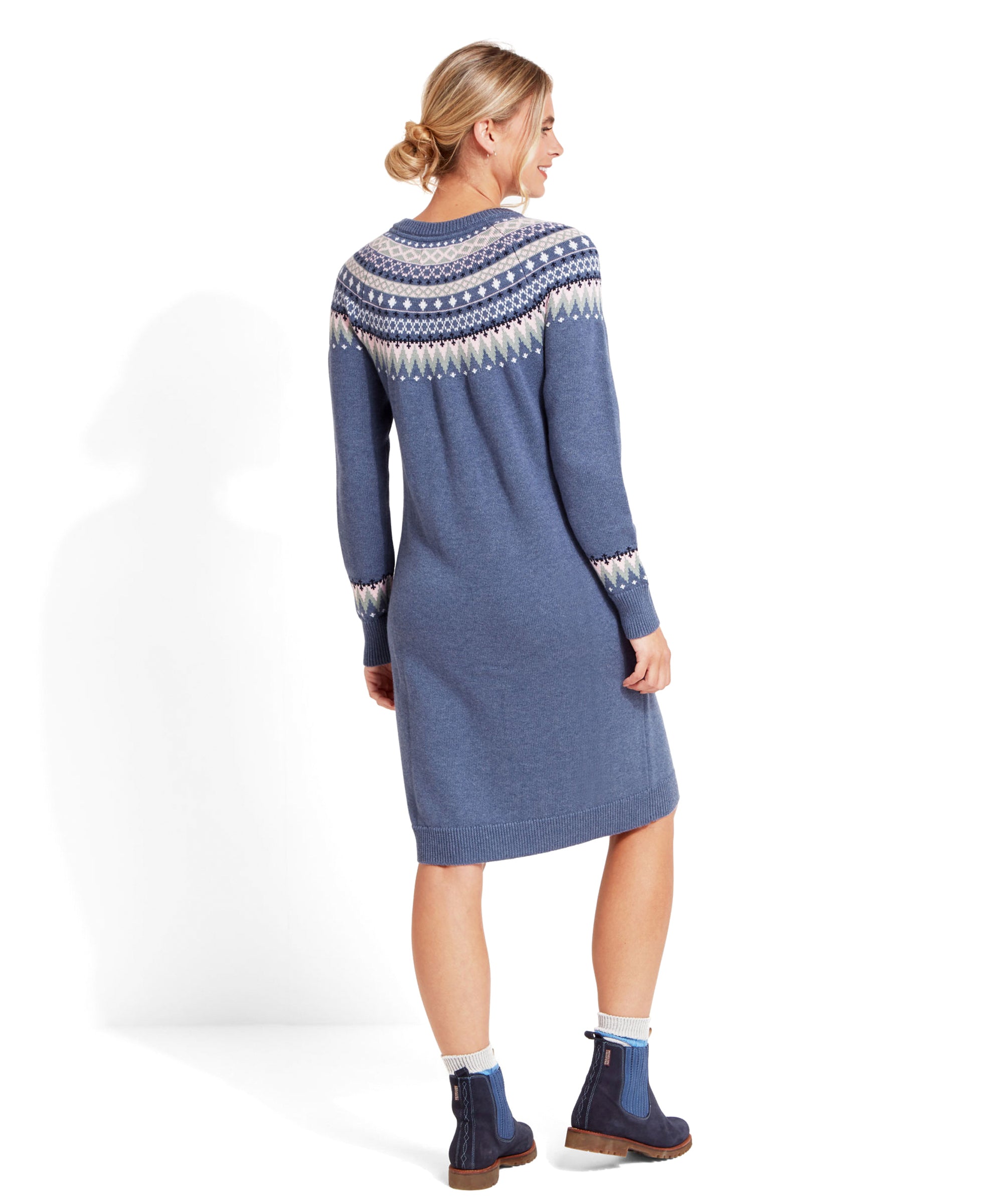 The back view of the same Schöffel Shetland Fairisle Dress for Women in Blue with the Fair Isle pattern. The detailed geometric pattern continues across the upper back and sleeves, showcasing the intricate design. The woman’s hair is styled in a loose, low bun, and she is looking over her shoulder with a smile. She pairs the dress with blue ankle boots and white socks.