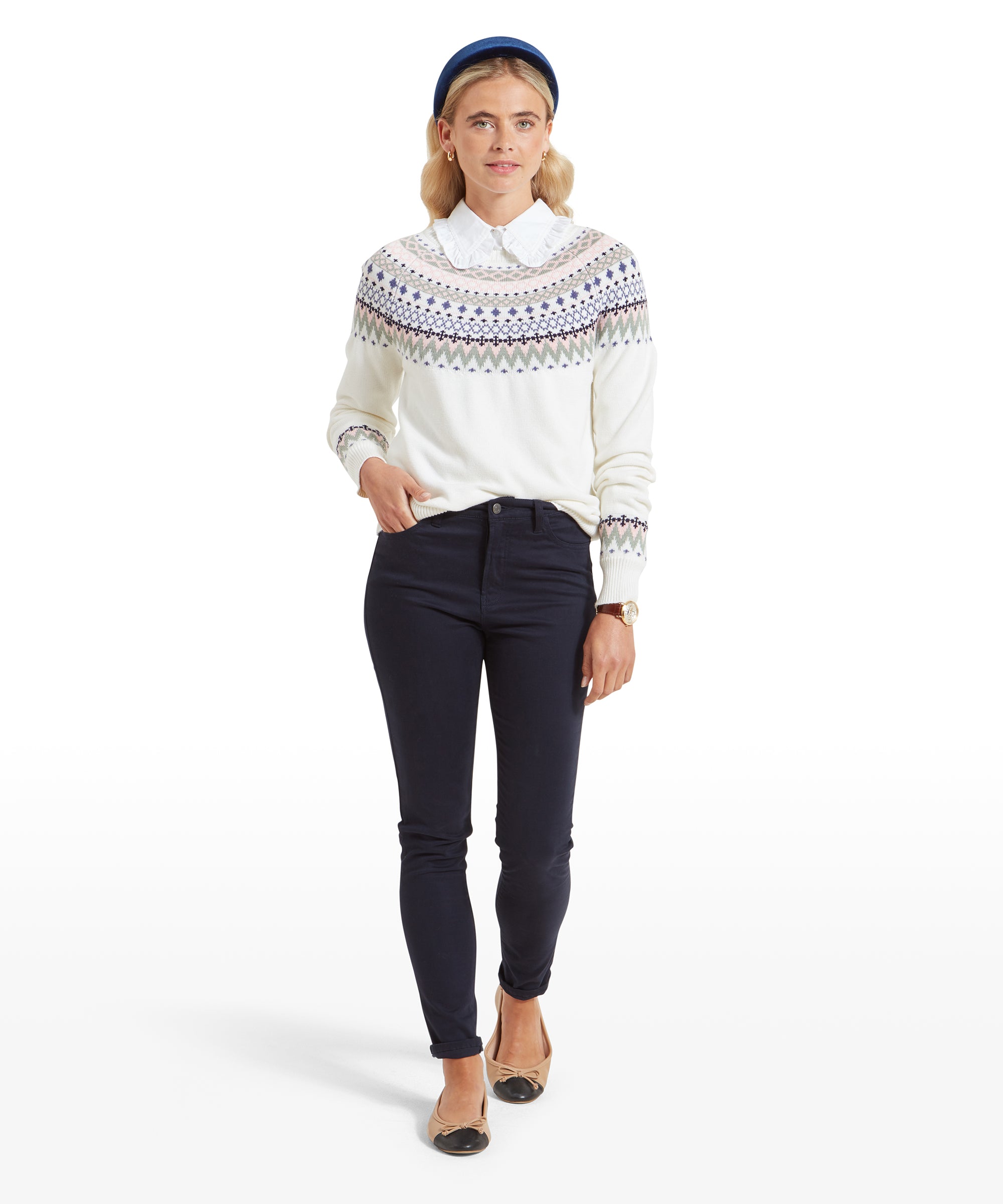 The woman is shown in full, wearing the same Schöffel Shetland Fairisle Jumper for Women in Pink Blue with a collared shirt underneath and dark navy pants. She completes her look with beige flats with black toe caps. She stands in a confident pose with her hands lightly clasped in front of her, projecting a classic and polished style.