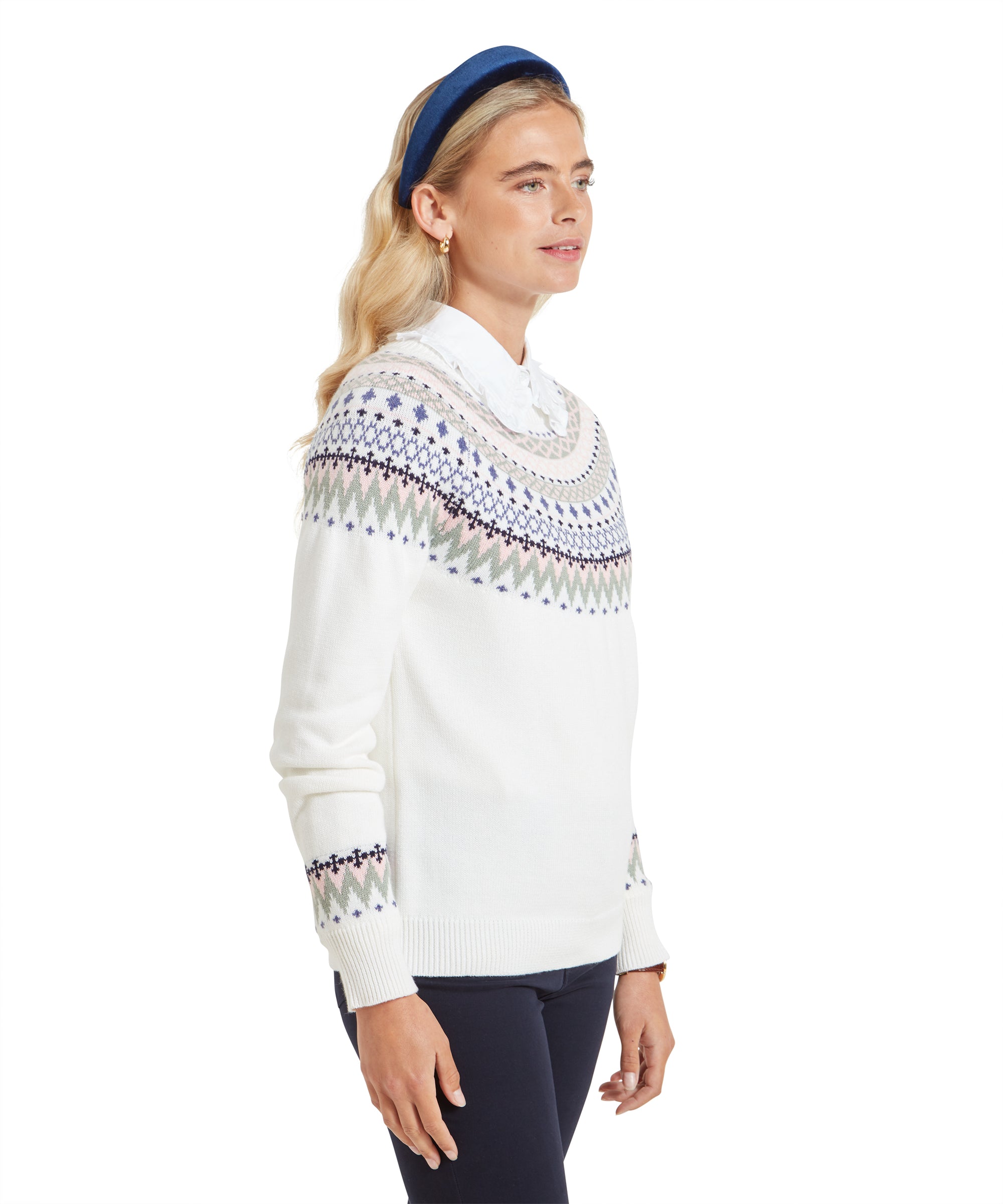 The woman is shown from a side angle, highlighting the Fair Isle pattern on her Schöffel Shetland Fairisle Jumper for Women in Pink Blue. Her blonde hair is styled in soft waves, and she wears a blue velvet headband. The outfit, paired with dark navy pants, has a refined and cosy appearance.