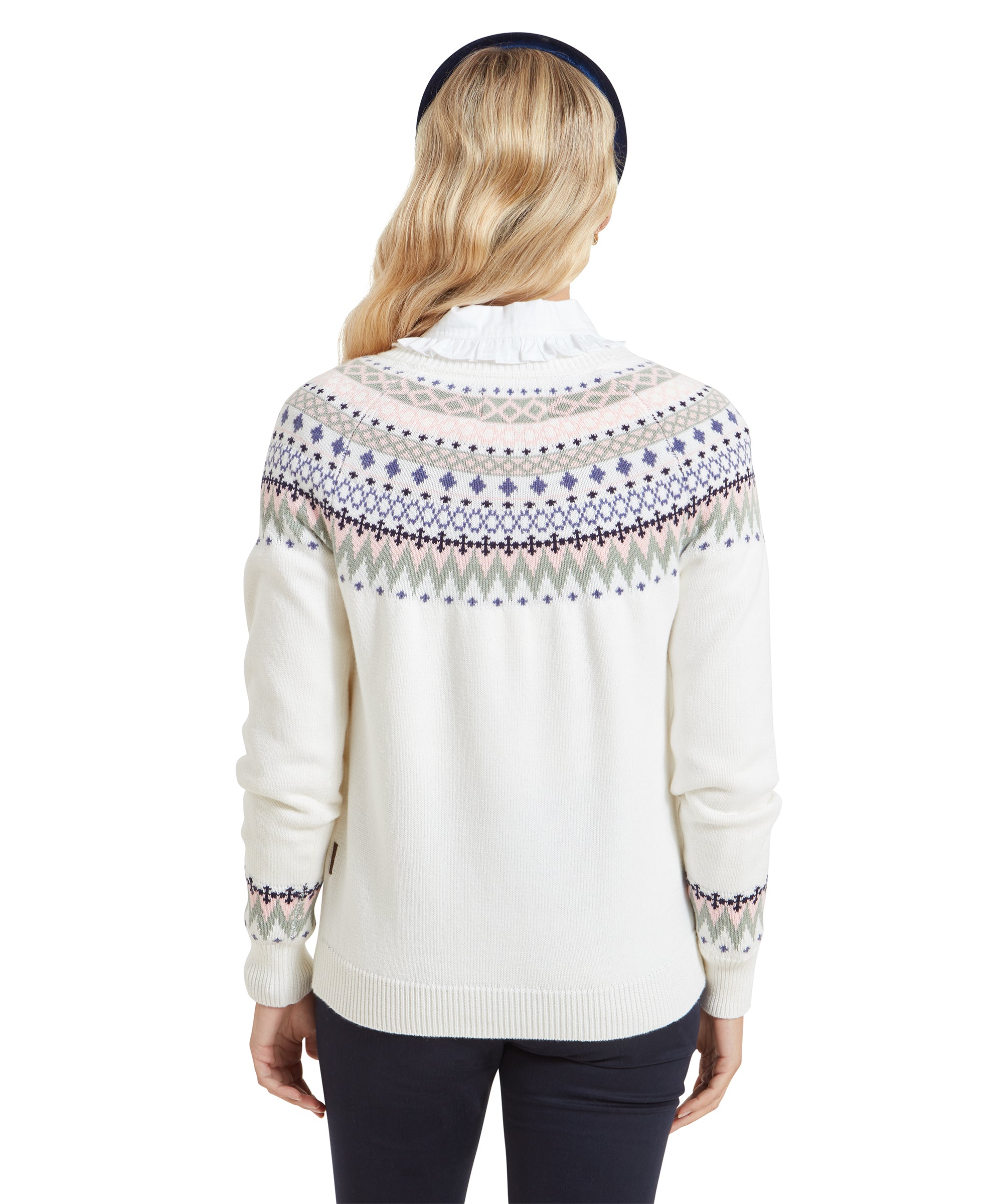 A back view of the woman showcases the intricate Fair Isle pattern across the upper back of the Schöffel Shetland Fairisle Jumper for Women in Pink Blue. Her blonde hair falls neatly down her back, and the sweater’s relaxed fit is evident. The colors of the pattern add a soft contrast to the crisp white base.