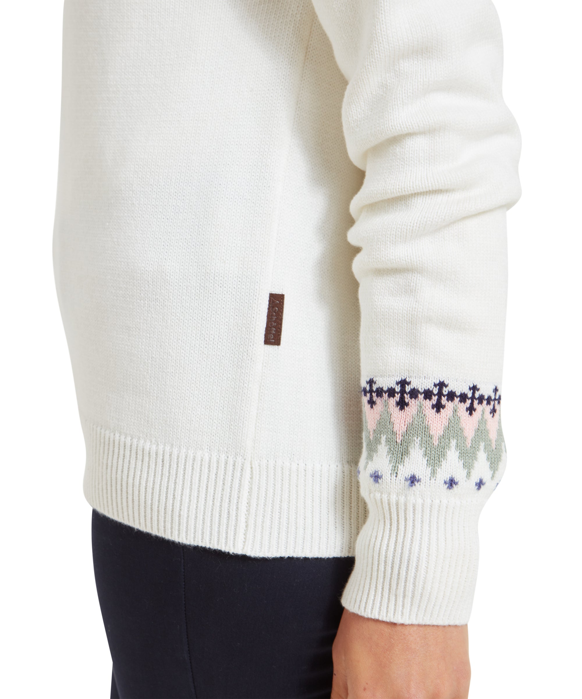 A close-up shot of the sleeve detail, emphasising the Fair Isle pattern at the cuff of the Schöffel Shetland Fairisle Jumper for Women in Pink Blue. The colors of blue, pink, and green are clearly visible against the white fabric, along with a small, brown fabric tag on the side seam.