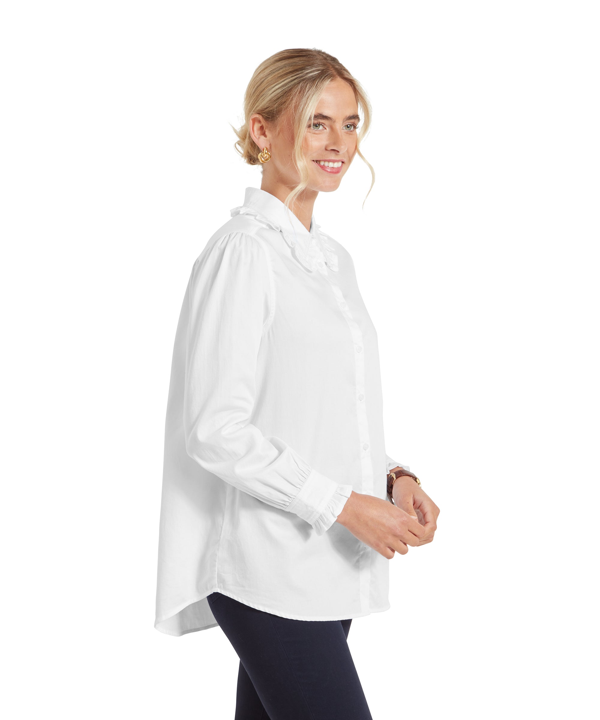 The woman is shown from the side, highlighting the loose fit of the Schöffel Somerleyton Shirt for Women in White. The shirt has a slight high-low hem, with the back being longer than the front. The ruffled collar and cuffs add a feminine touch to the shirt.