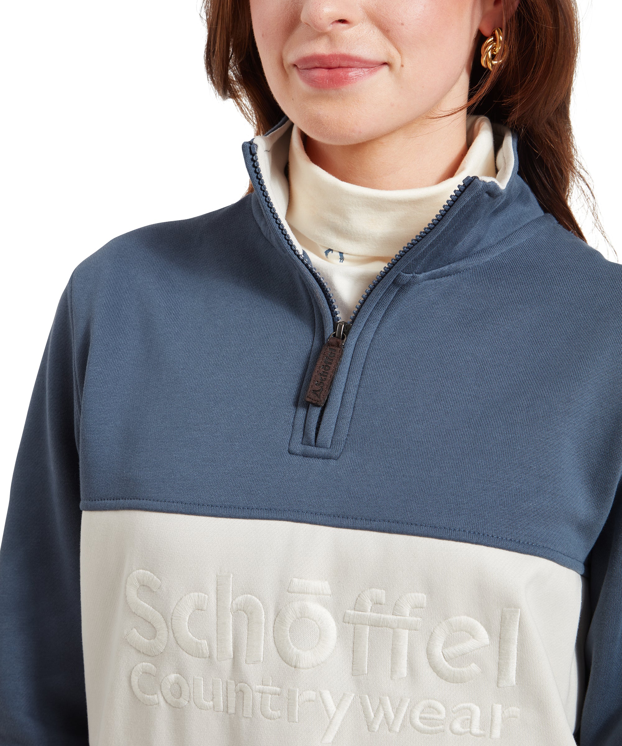 A close-up of the woman's upper body, focusing on the quarter-zip of the Schöffel St Agnus Sweatshirt for Women in Navy. The embroidered "Schöffel Countrywear" logo is clearly visible on the cream panel across the chest, and the zipper pull has a small leather detail.