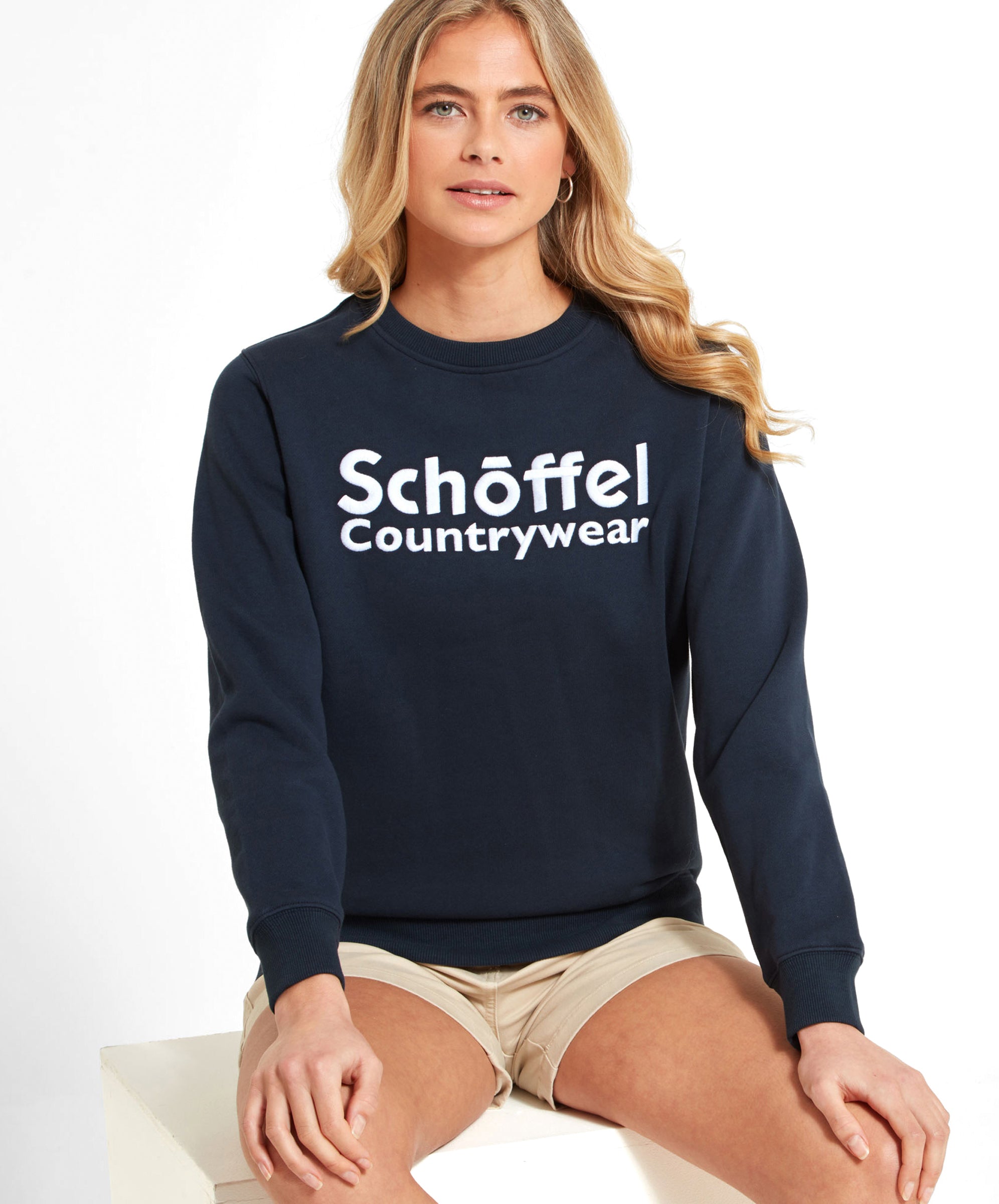 A woman is wearing the Schöffel St Helier Sweatshirt for Women in Navy. The sweatshirt features the Schöffel Countrywear logo prominently on the front. She is sitting down with her hands resting on her thighs, wearing beige shorts.