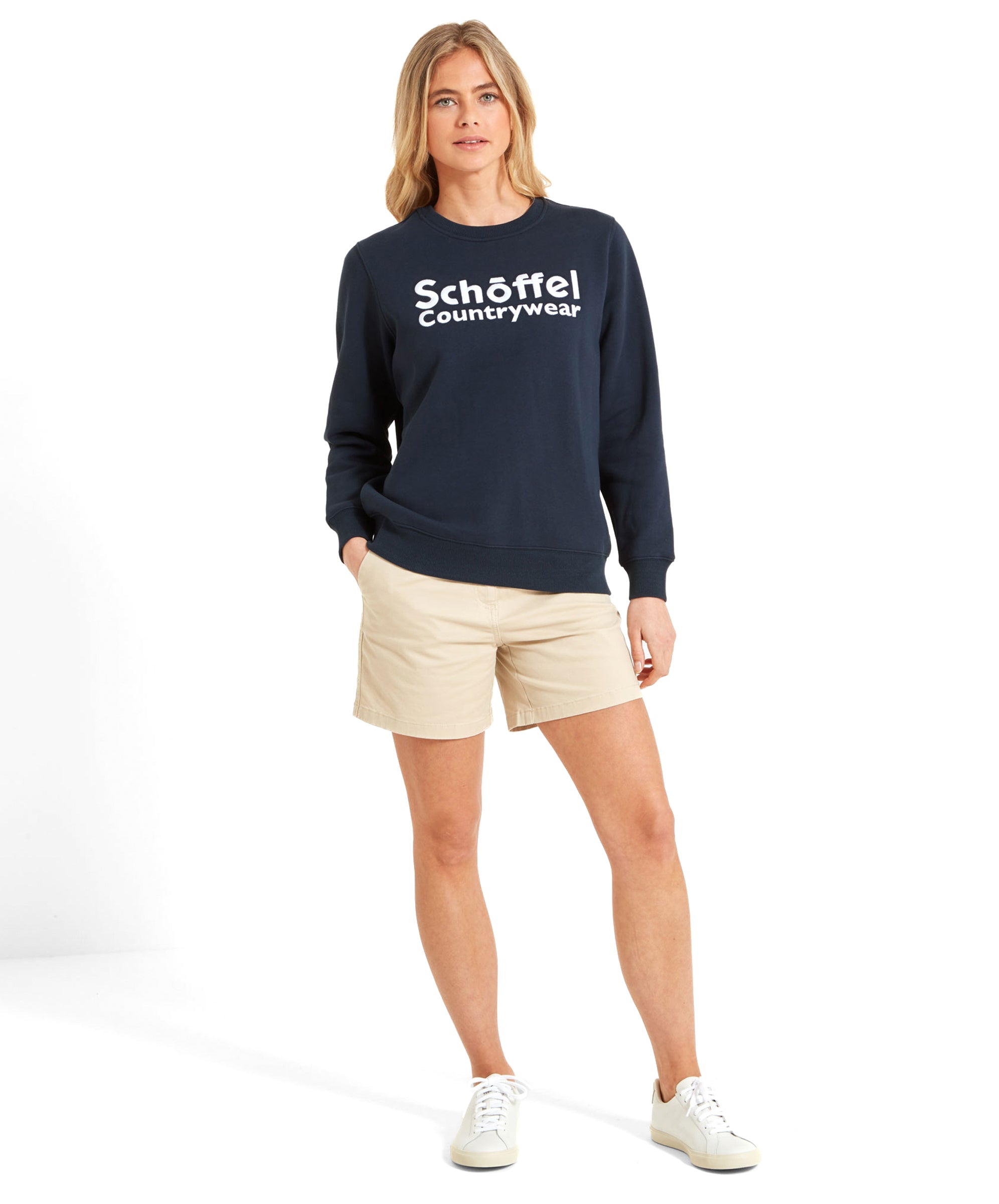 The woman is standing, modeling the Schöffel St Helier Sweatshirt for Women in Navy. She has her hands in the pockets of her beige shorts, pairing the outfit with white trainers.