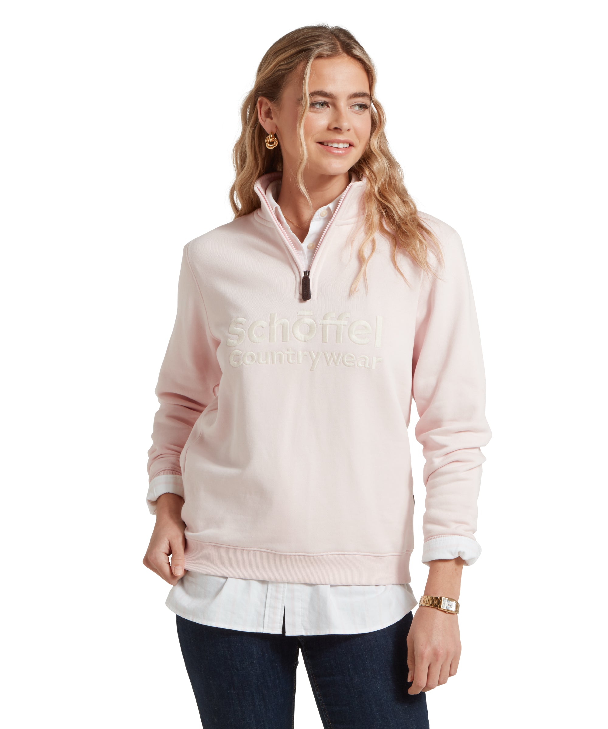 A woman wearing a Schöffel St Issey Sweatshirt for Women in Light Pink with "Schöffel Countrywear" embroidered in matching light beige across the chest. She is wearing dark blue jeans, and a white collared shirt underneath the sweatshirt. She looks slightly to her left with a relaxed expression.