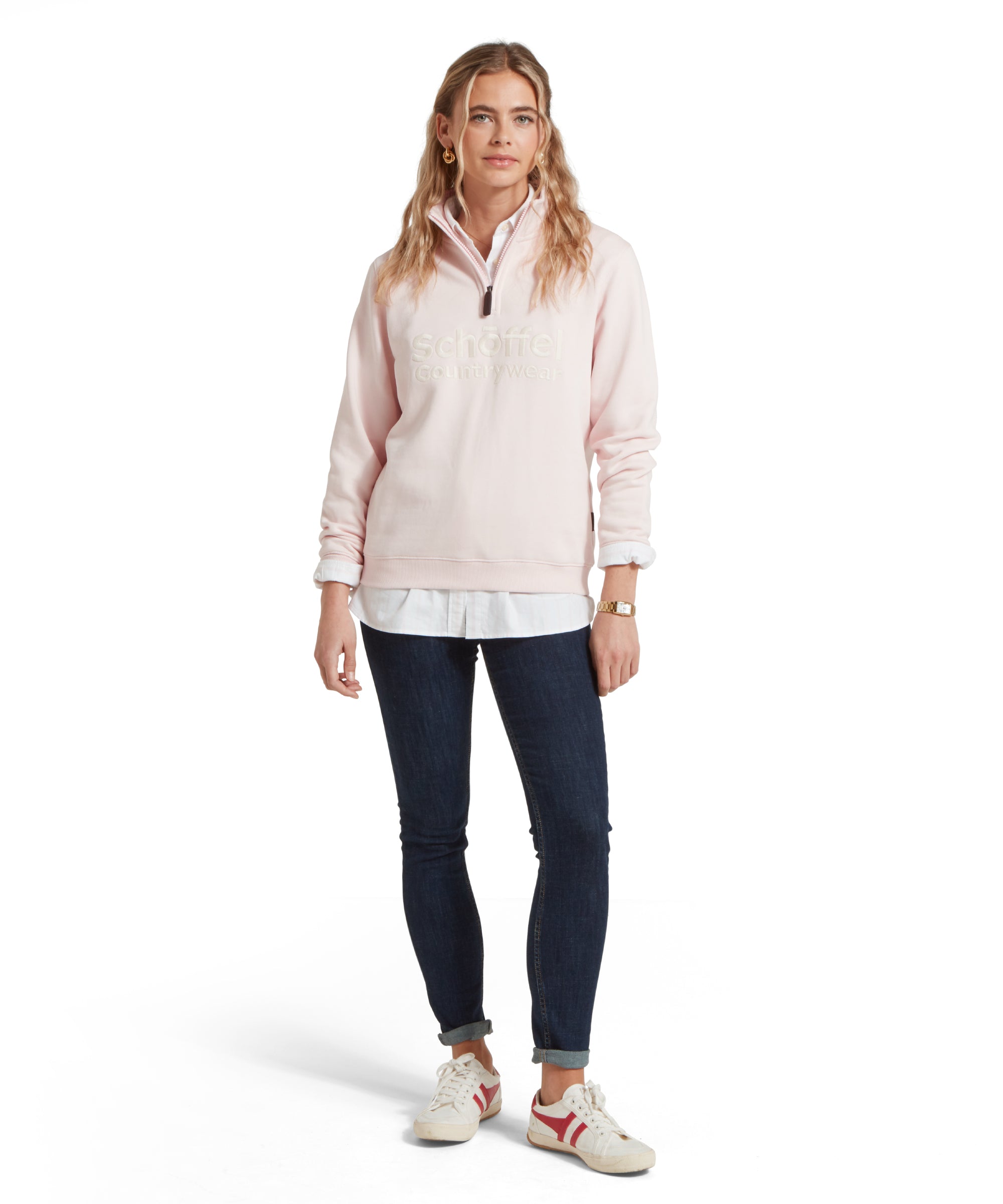 A full-body image of a woman dressed in a Schöffel St Issey Sweatshirt for Women in Light Pink with "Schöffel Countrywear" embroidered across the chest, dark blue jeans, and white trainers. She has her blonde hair half tied back and is looking towards the camera with a slight smile.