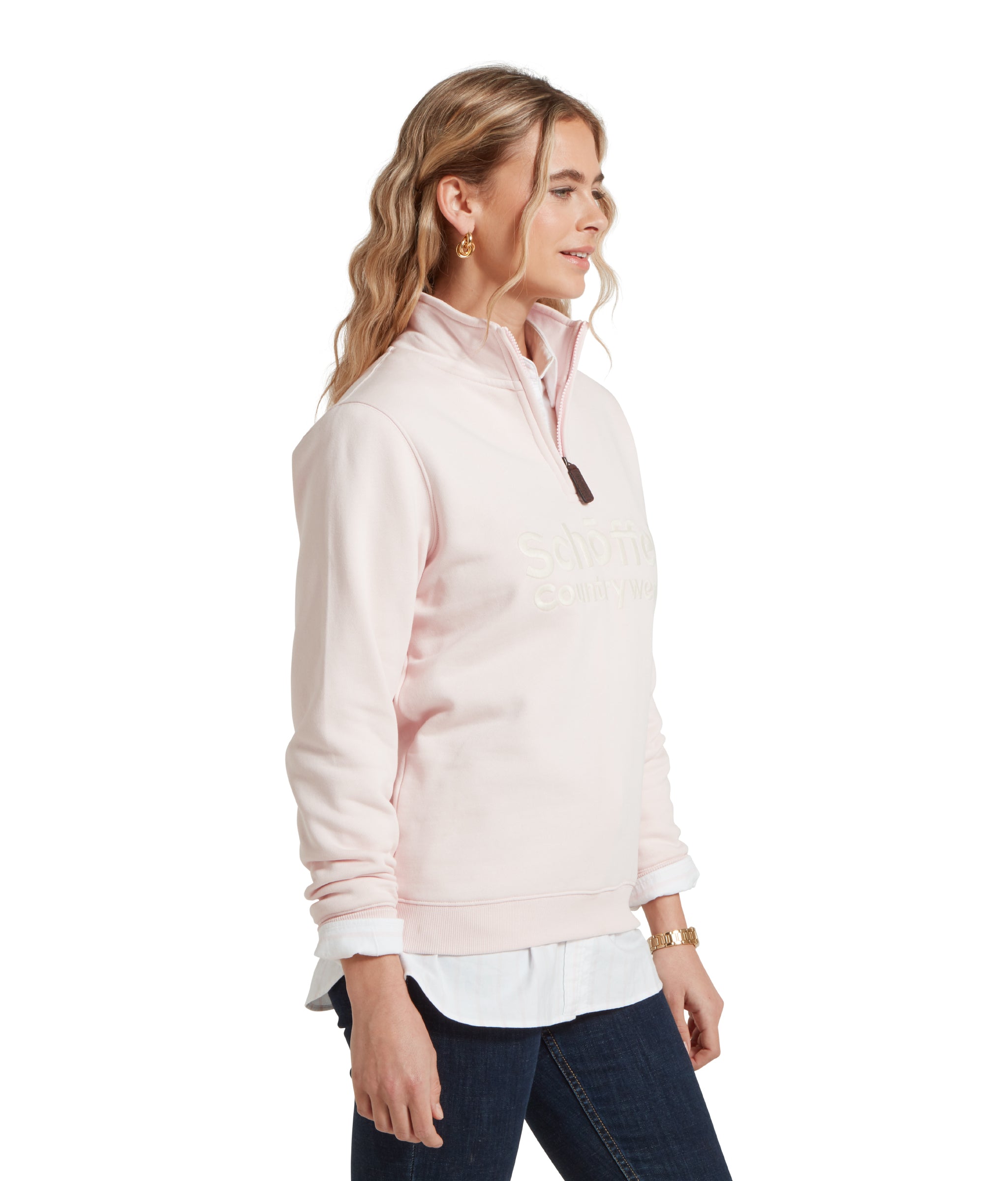 A side profile of a woman standing and facing her left, wearing a Schöffel St Issey Sweatshirt for Women in Light Pink with "Schöffel Countrywear" embroidered across the chest, dark blue jeans, and a white collared shirt underneath.