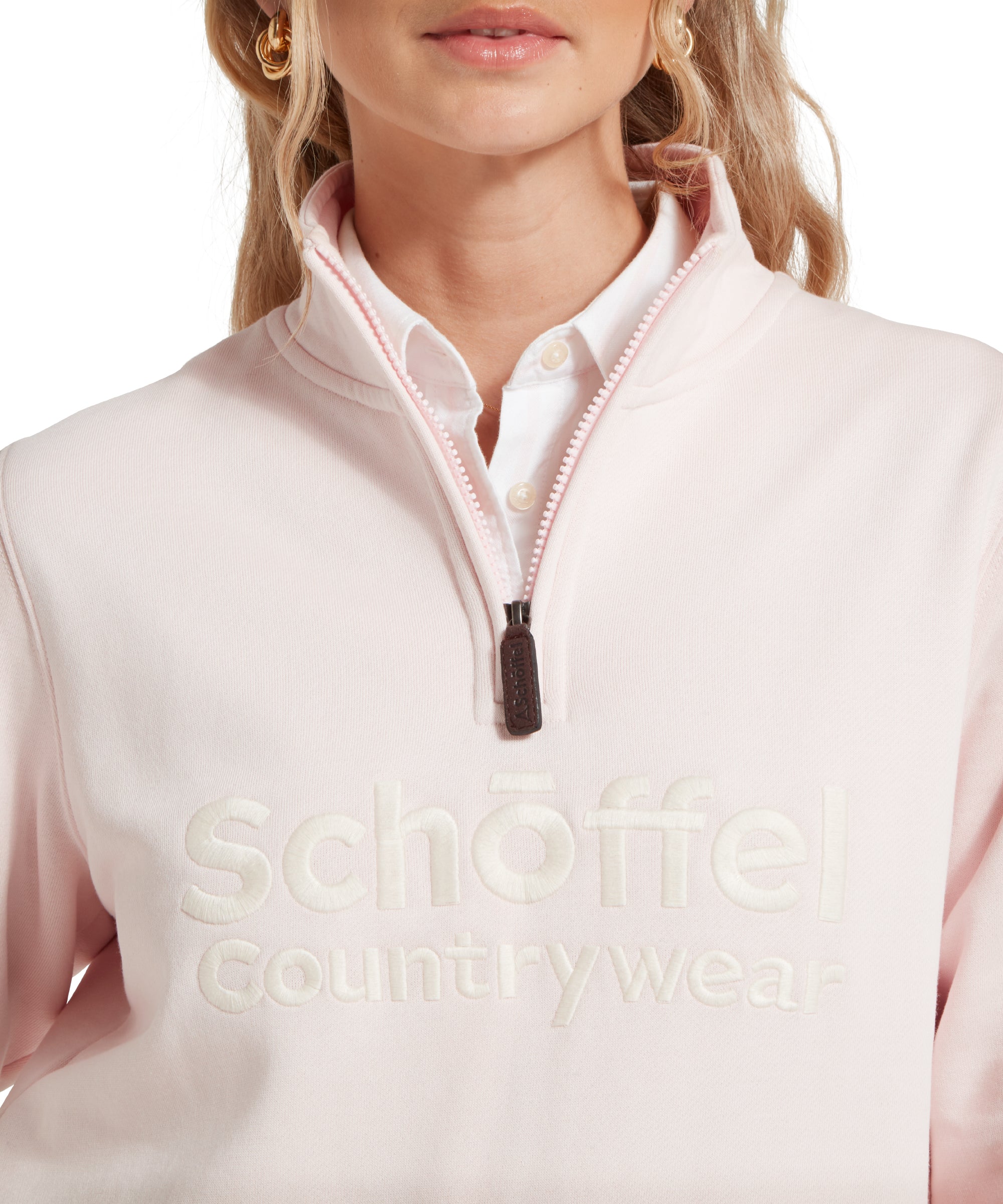 A close-up image of the upper part of the Schöffel St Issey Sweatshirt for Women in Light Pink, highlighting the embroidered "Schöffel Countrywear" logo across the chest. The image also shows the half-zip detail with a leather pull tab and the white collared shirt peeking out from underneath the sweatshirt.