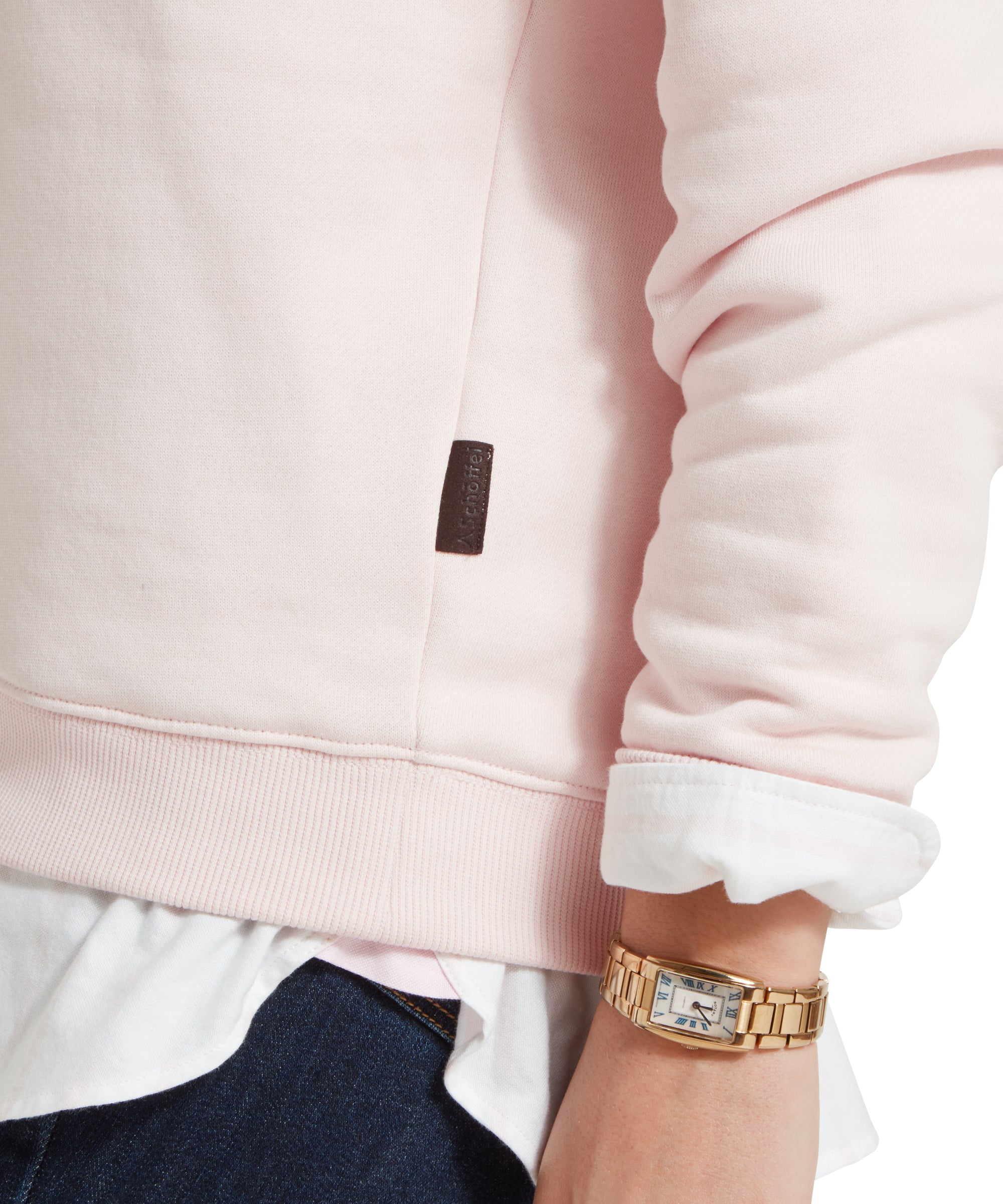 A close-up shot focusing on the lower left side of the Schöffel St Issey Sweatshirt for Womens in Light Pink, showing the ribbed hem and a small brown tag with the brand’s name. The sleeve is rolled up slightly to reveal the white cuff of the shirt underneath. The edge of dark blue jeans is visible.