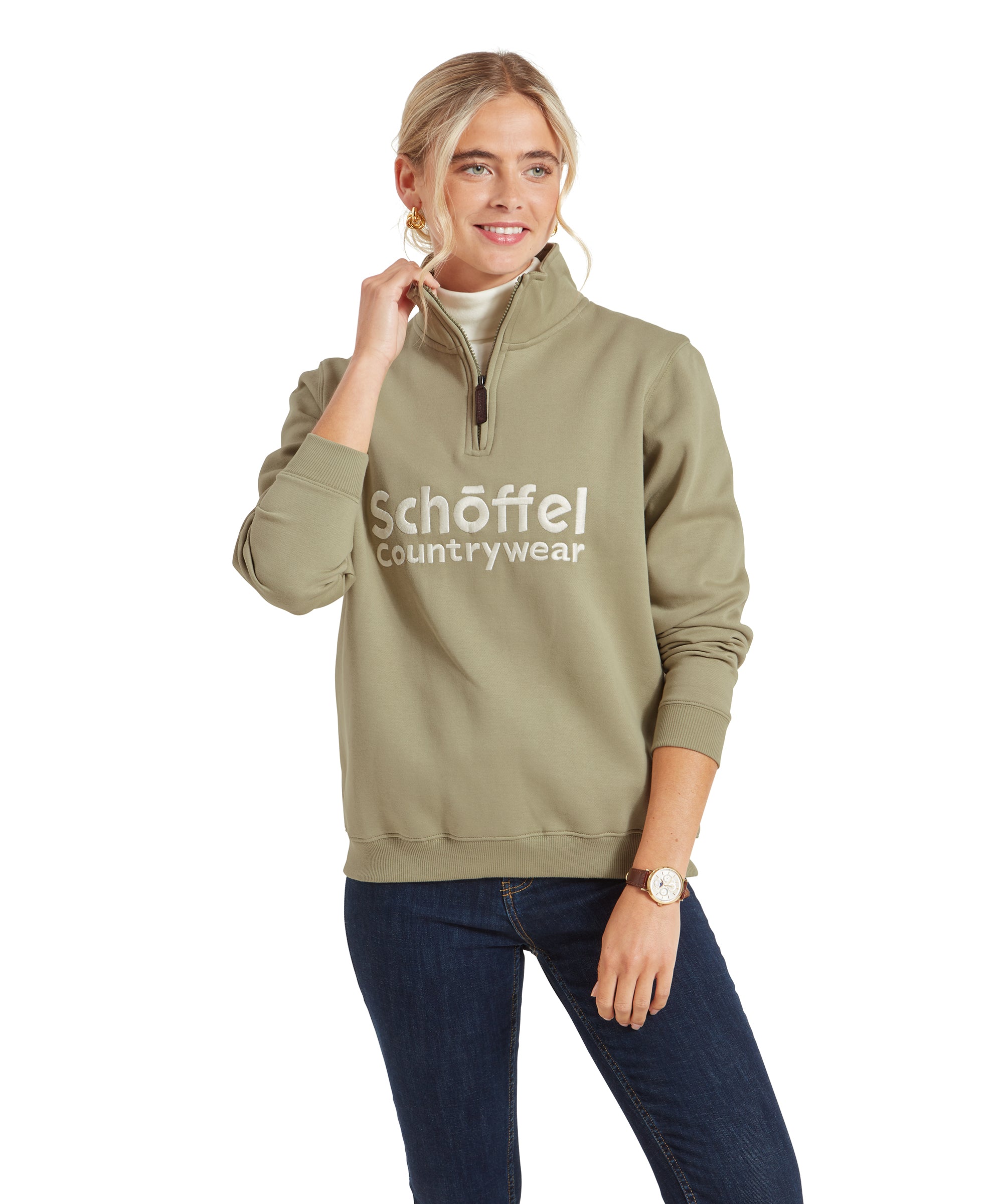 A woman is standing, modeling a Schöffel St Issey Sweatshirt for Womens in Green with a half-zip collar. She is smiling slightly, with one hand gently pulling at the zipper. The sweatshirt has the "Schöffel Countrywear" logo prominently embroidered in white across the chest. She is wearing dark jeans and has her hair pulled back into a loose ponytail. She also has gold hoop earrings and a watch on her left wrist.