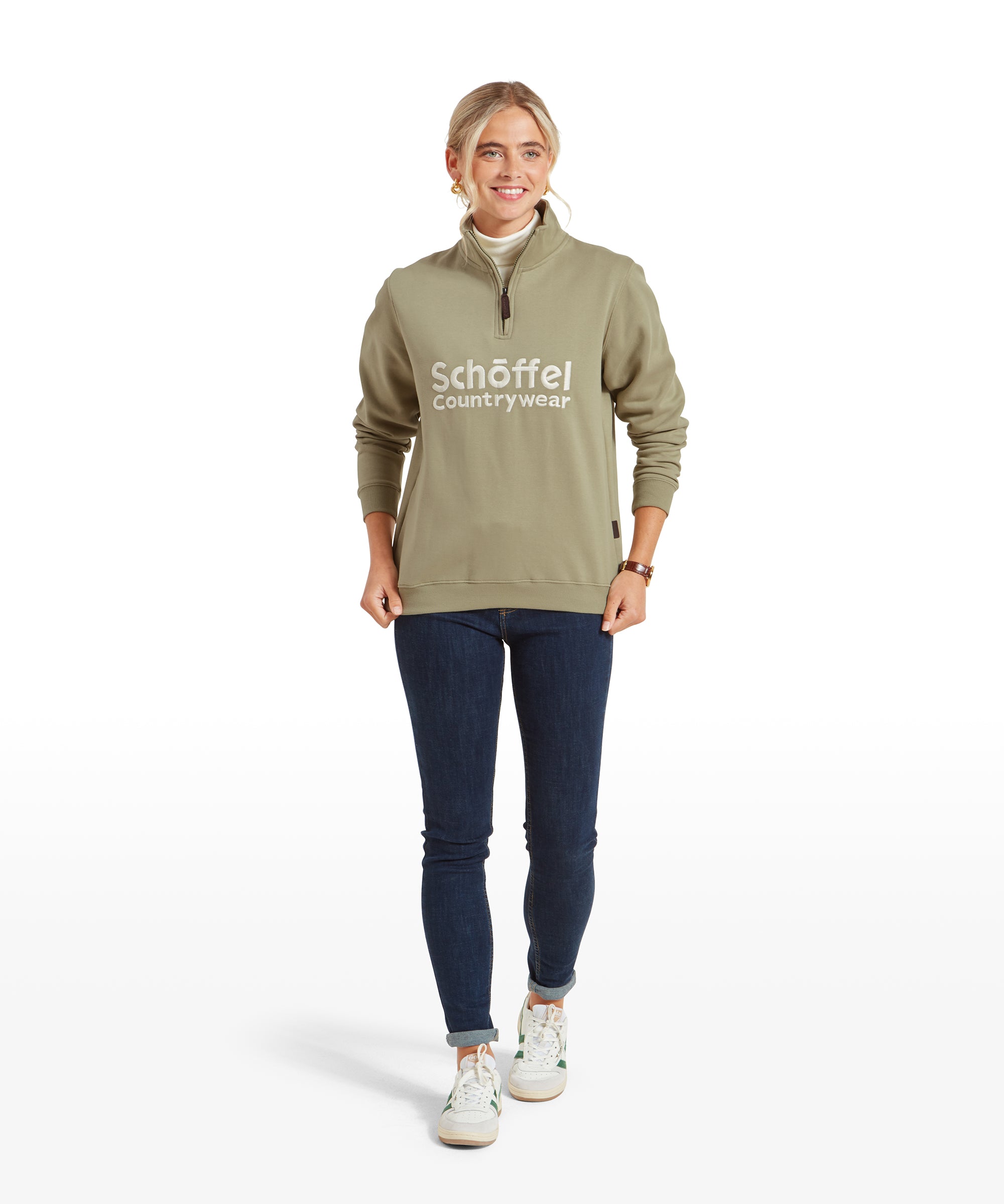 The same model is shown in a full-body view, walking forward with her arms slightly bent. She is wearing the Schöffel St Issey Sweatshirt for Womens in Green with the logo across the chest, paired with dark skinny jeans and white sneakers. Her expression is relaxed and confident.