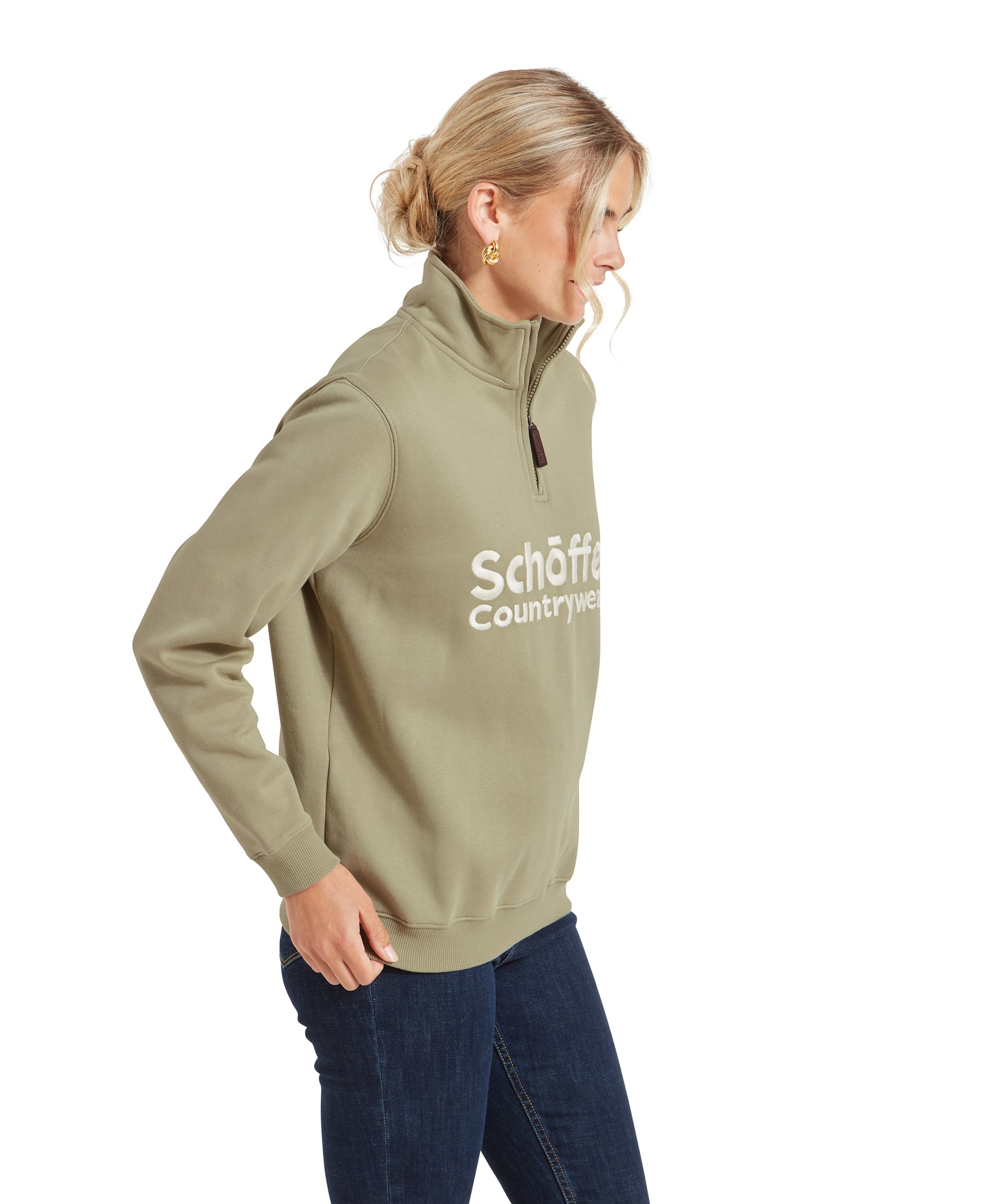 A side profile of the model wearing the Schöffel St Issey Sweatshirt for Womens in Green. She is looking down slightly, with her left arm extended down and her right arm relaxed by her side. The sweatshirt's zip is partially up, showing a bit of the white turtleneck underneath.