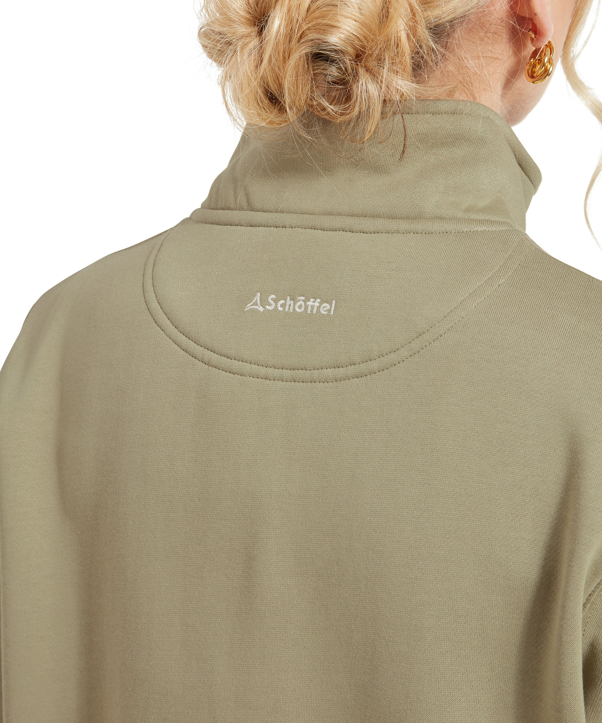 A close-up shot of the back of the Schöffel St Issey Sweatshirt for Womens in Green, focusing on the embroidered Schöffel logo just below the collar. The stitching detail and fabric texture are visible, with the model's blonde hair pulled back into a neat bun.