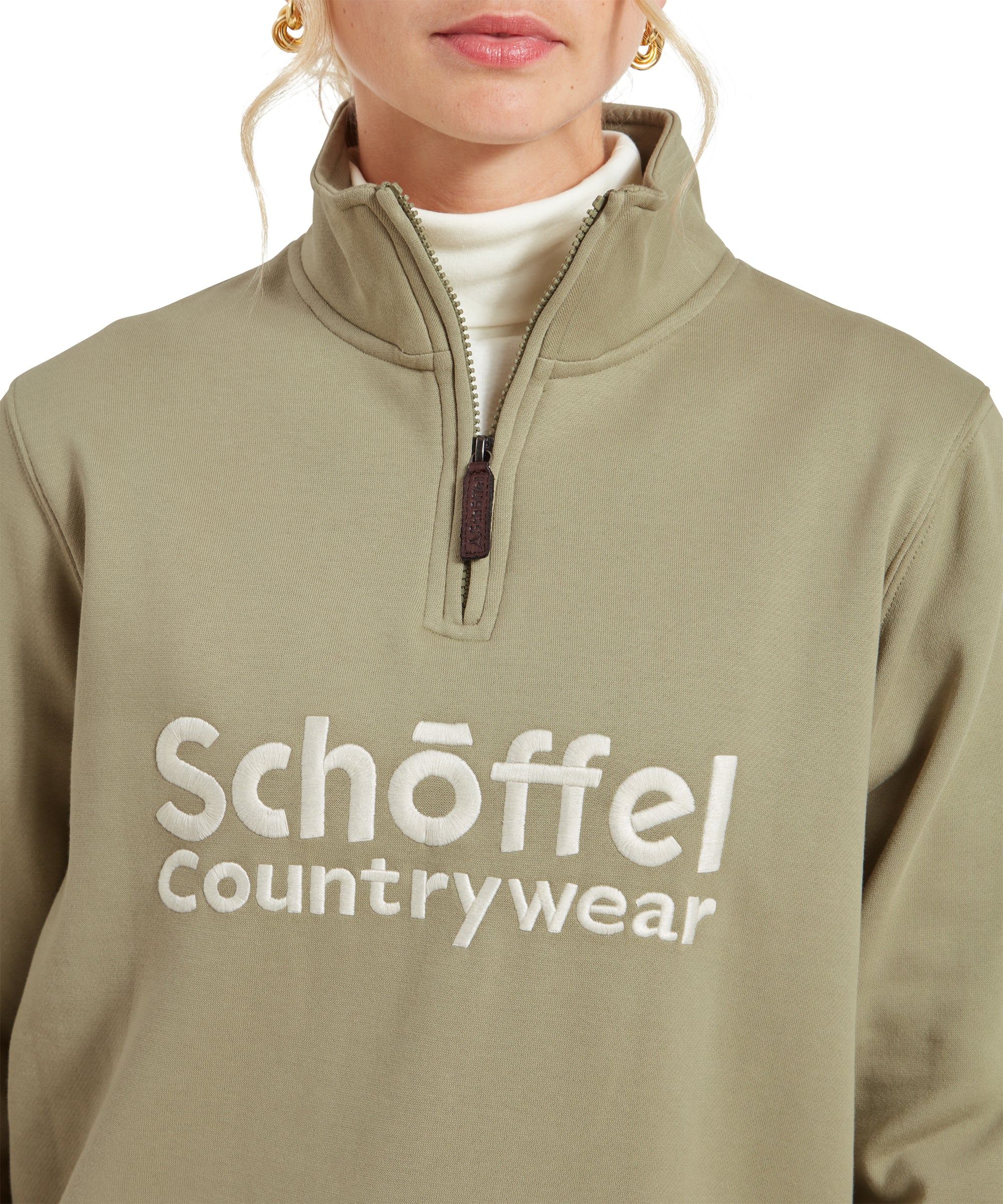 A close-up view of the front of the Schöffel St Issey Sweatshirt for Womens in Green, highlighting the half-zip collar and the embroidered Schöffel Countrywear logo on the chest. The model’s face is partially visible, and she is wearing a white turtleneck underneath the sweatshirt, paired with gold hoop earrings.