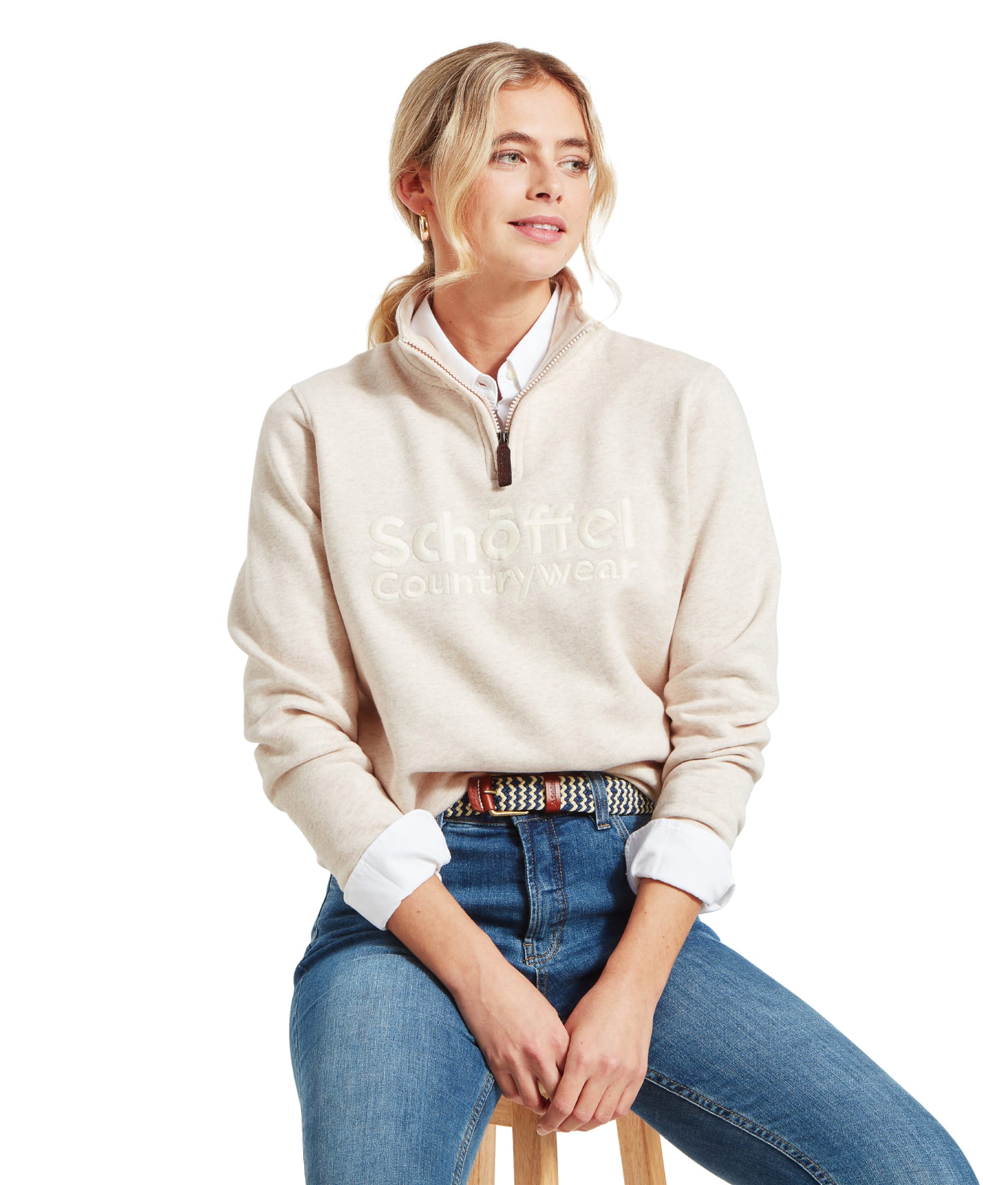 A woman sitting on a wooden stool, wearing a Schöffel St Issey Sweatshirt for Womens in Brown with "Schöffel Countrywear" embroidered in matching light beige across the chest. She has her blonde hair tied back in a low ponytail, is wearing medium blue jeans, and a white collared shirt underneath the sweatshirt. She looks slightly to her left with a relaxed expression.