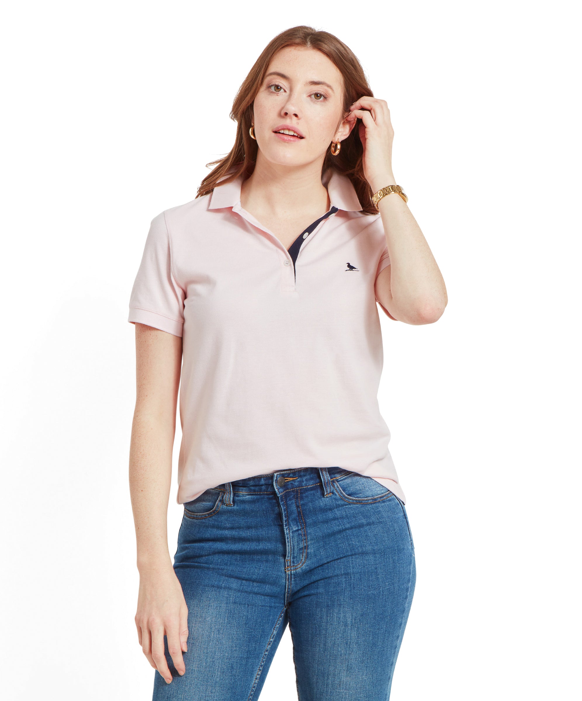 A model wearing a Schöffel Women's St Ives Polo Shirt for Women in Pale Pink with blue jeans, accessorised with gold hoop earrings and a gold watch.