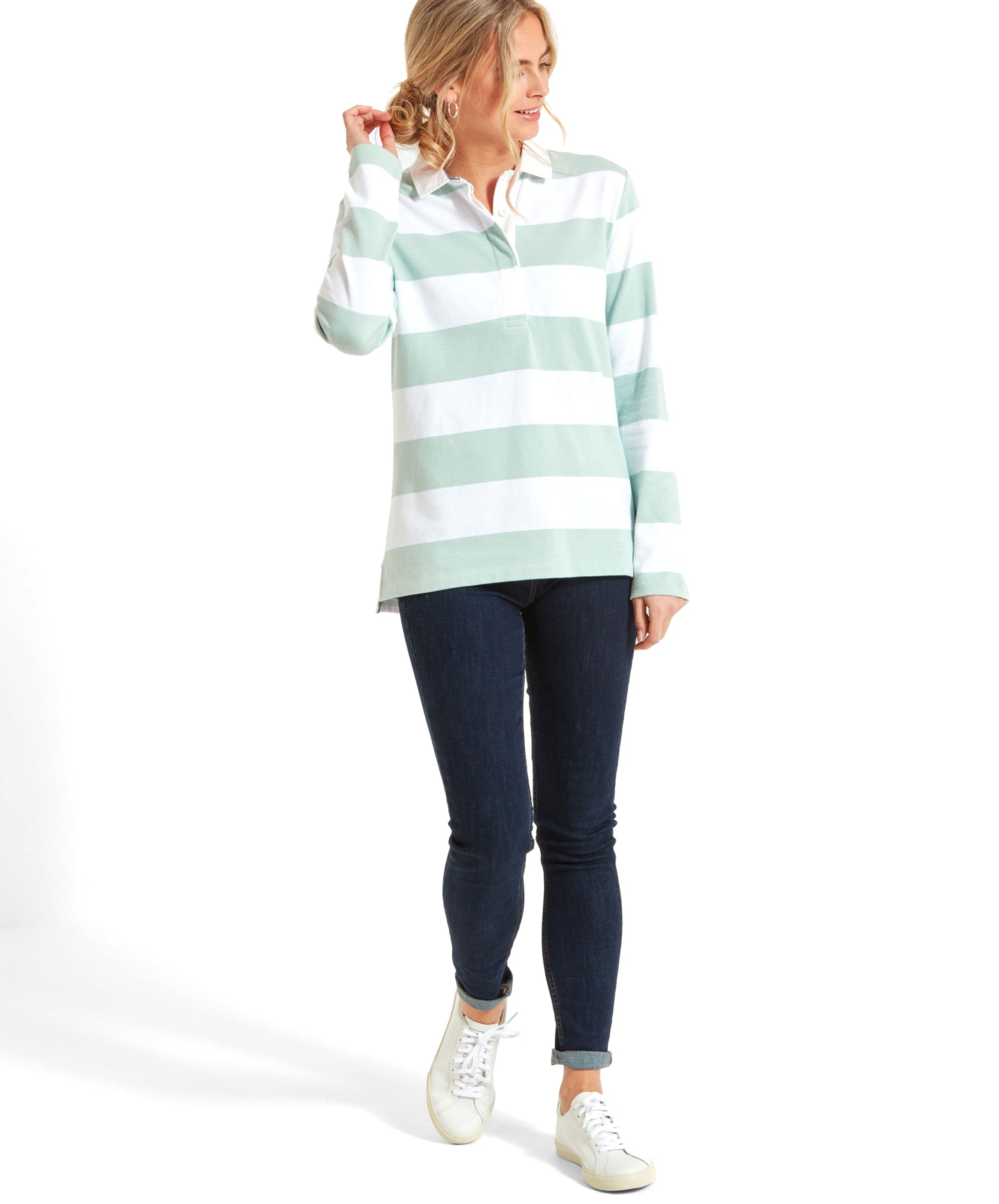 A full-body view of the woman wearing the Schöffel St Mawgan Rugby Shirt for Women in Green. The shirt has wide green and white stripes and a white collar. The model is looking to the side with a smile, and she has one hand gently touching her neck.