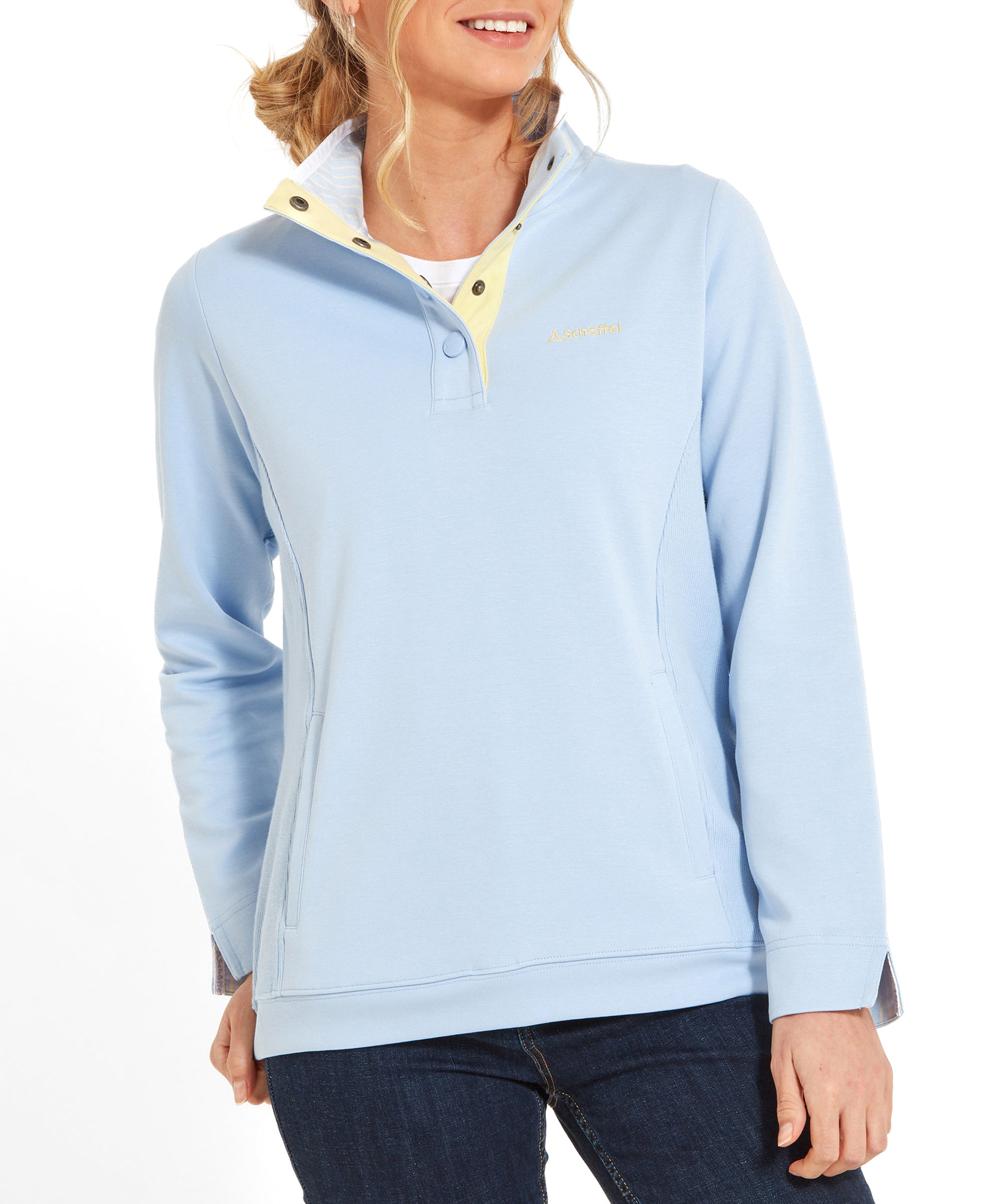 A woman wearing a Schöffel Steephill Cove Sweatshirt for Women in Blue with a high collar and snap buttons. The sweatshirt features the Schoffel logo in yellow on the left chest. She is smiling and her blonde hair is tied back in a ponytail. She pairs the sweatshirt with dark jeans.