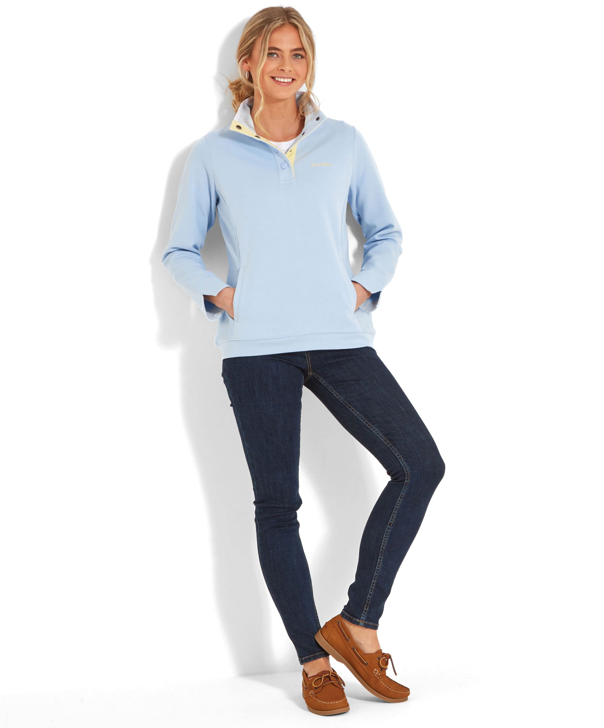 Full body view of the woman standing against a white background, wearing the same Schöffel Steephill Cove Sweatshirt for Women in Blue. She has her hands in the pockets of her dark jeans and is wearing brown loafers. She is smiling and leaning slightly to one side.