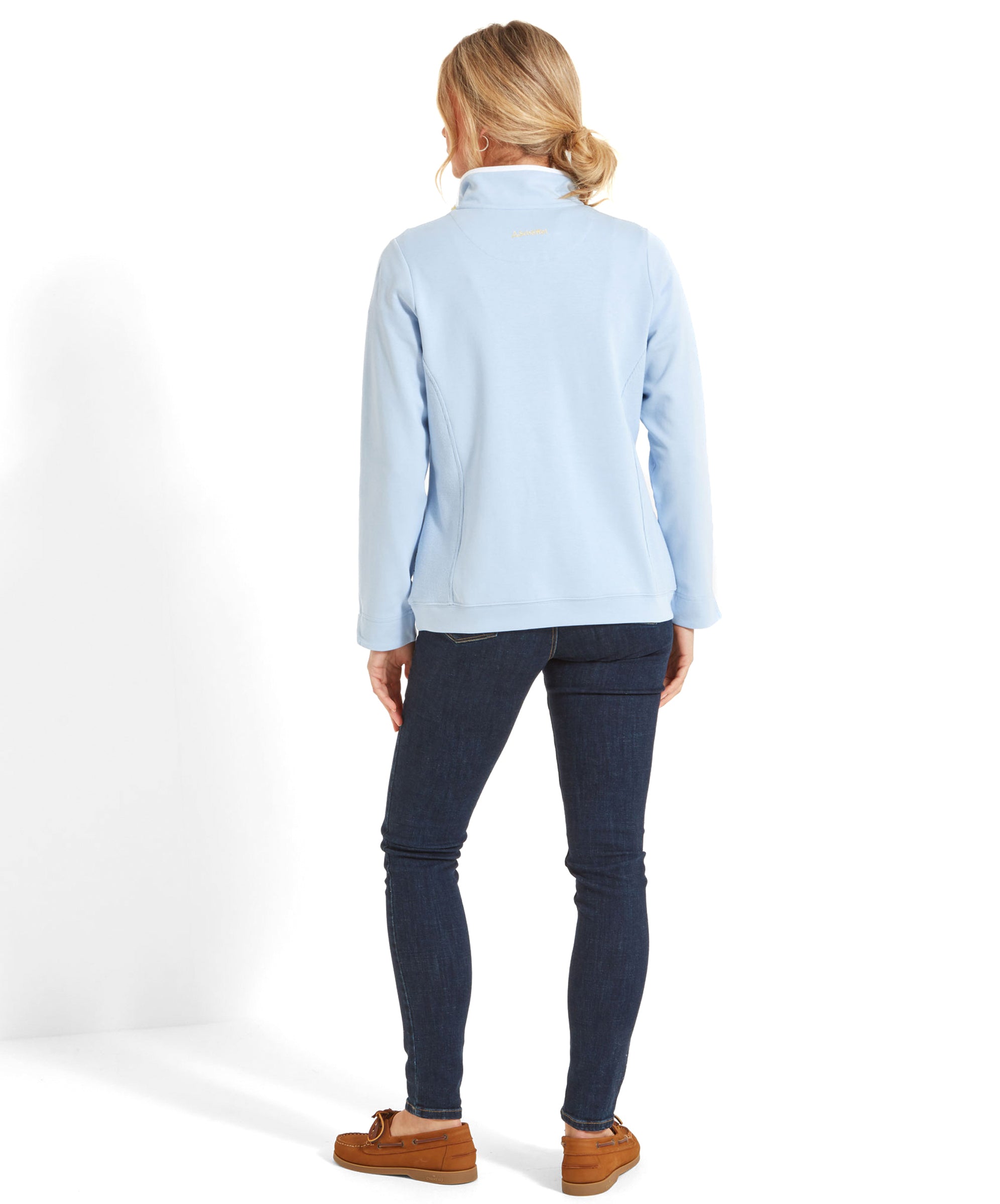 Rear view of the woman showcasing the back of the Schöffel Steephill Cove Sweatshirt for Women in Blue. The sweatshirt has a simple, clean design with the Schoffel logo in yellow near the collar. She is wearing dark jeans and brown loafers, standing with her back to the camera.