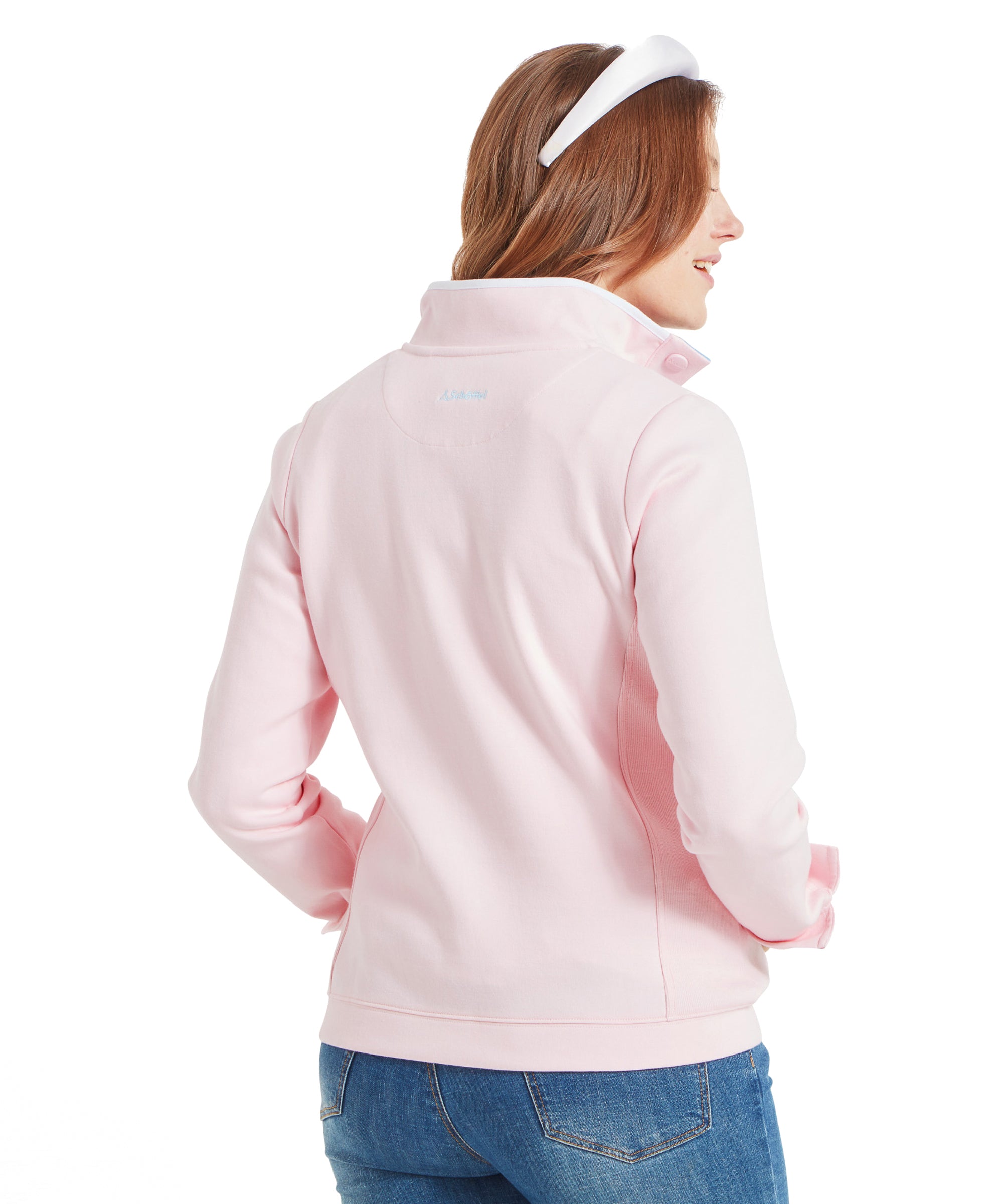 Back view of the woman wearing the Schöffel Steephill Cove Sweatshirt for Women in Pink. The sweatshirt has a high collar and is paired with blue jeans. The woman's hair is styled with a white headband.