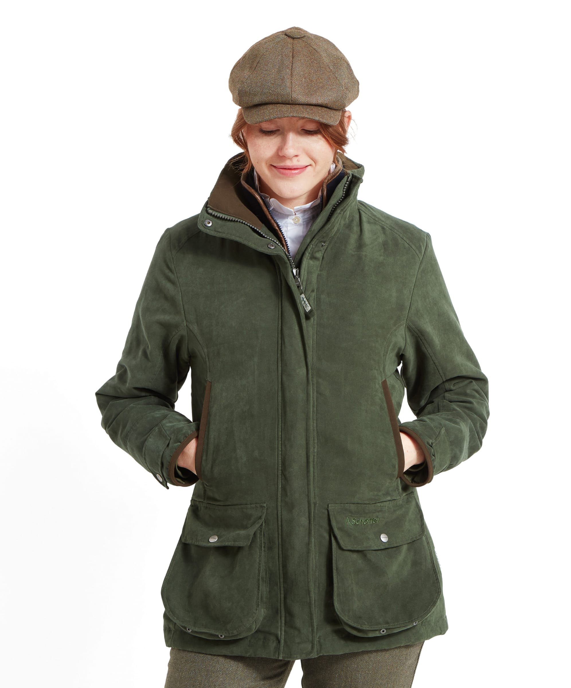 A woman is wearing an Schöffel Teal Shooting Coat for Women in Green with a high collar and large front pockets, paired with a tweed flat cap. She is standing with her hands in the jacket pockets, looking down with a slight smile. The jacket has a smooth suede-like finish and a structured, fitted design.