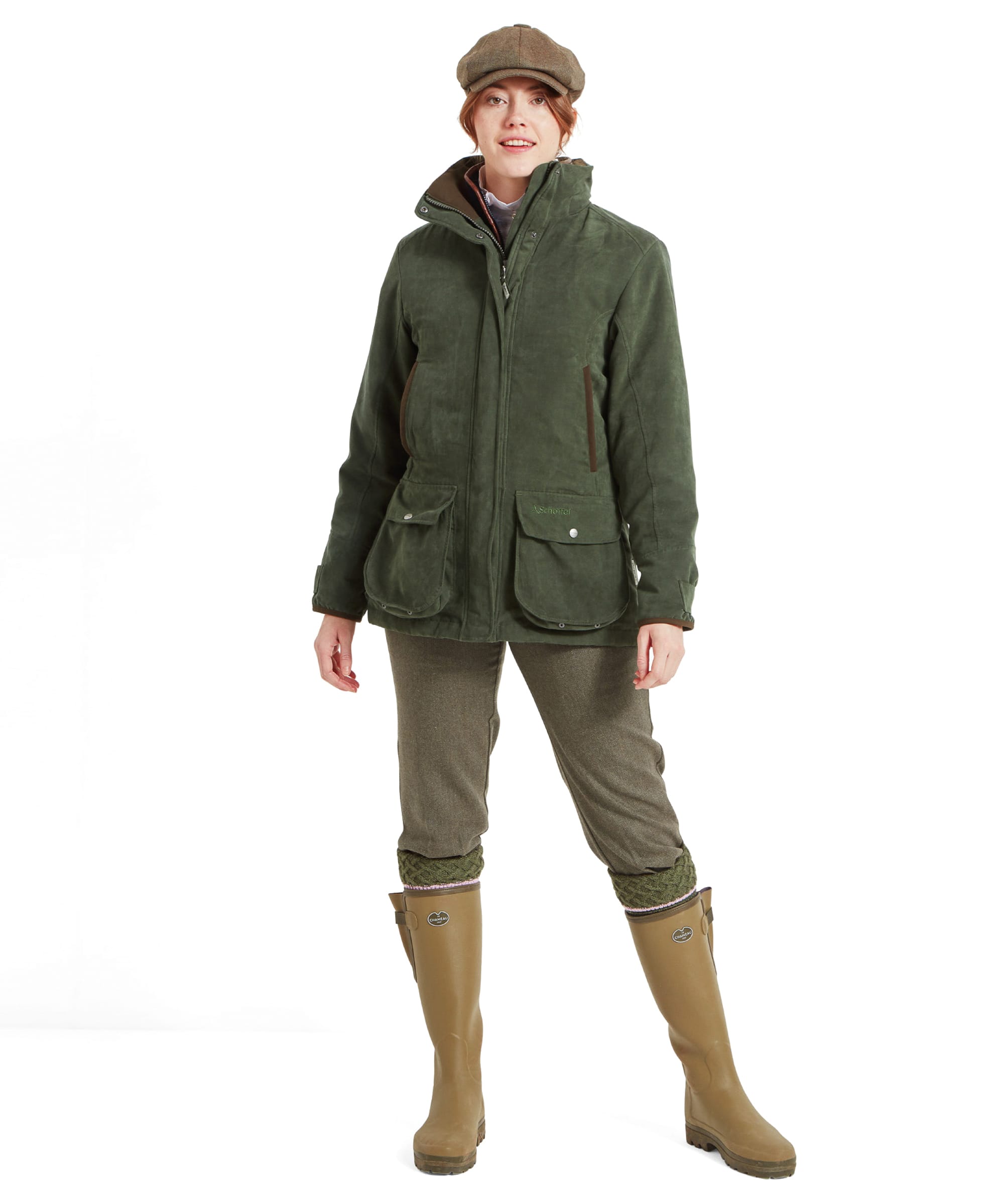 A full-body shot of the woman wearing the Schöffel Teal Shooting Coat for Women in Green, olive-green pants, and knee-high tan rubber boots. She stands confidently with her hands in her pockets, facing forward. The jacket’s high collar is fully zipped up, and the tweed flat cap complements the outfit.