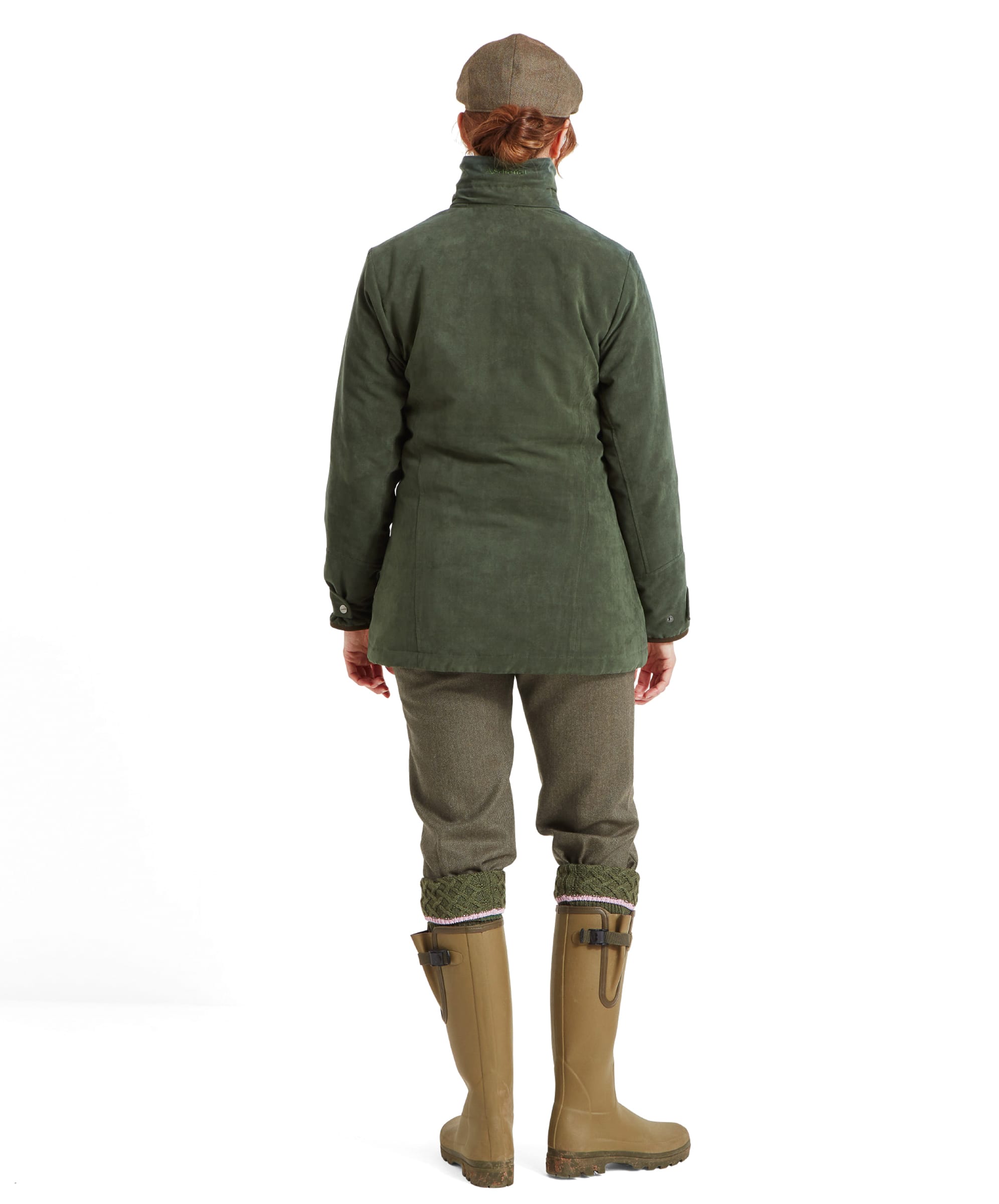 A rear view of the woman, highlighting the back of the Schöffel Teal Shooting Coat for Women in Green. The jacket’s collar is raised, and the back of the jacket is smooth and simple. The woman is wearing olive-green pants and tan rubber boots, with the back of the tweed flat cap visible.