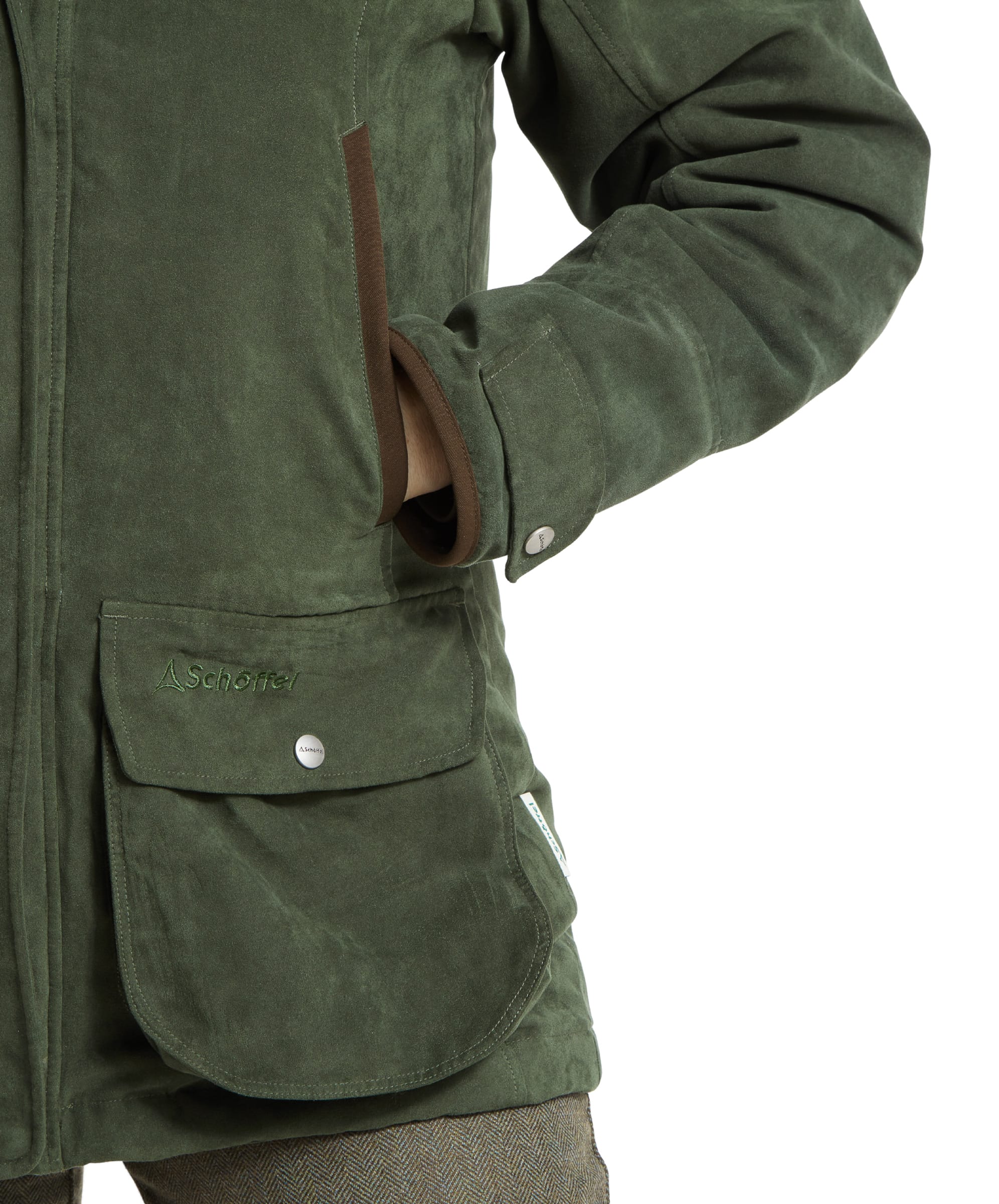 A close-up of the left front pocket of the Schöffel Teal Shooting Coat for Women in Green, with the woman’s hand tucked inside. The pocket flap is secured with a snap button and features subtle brown trim along the edge. The suede-like fabric and craftsmanship are clearly visible.