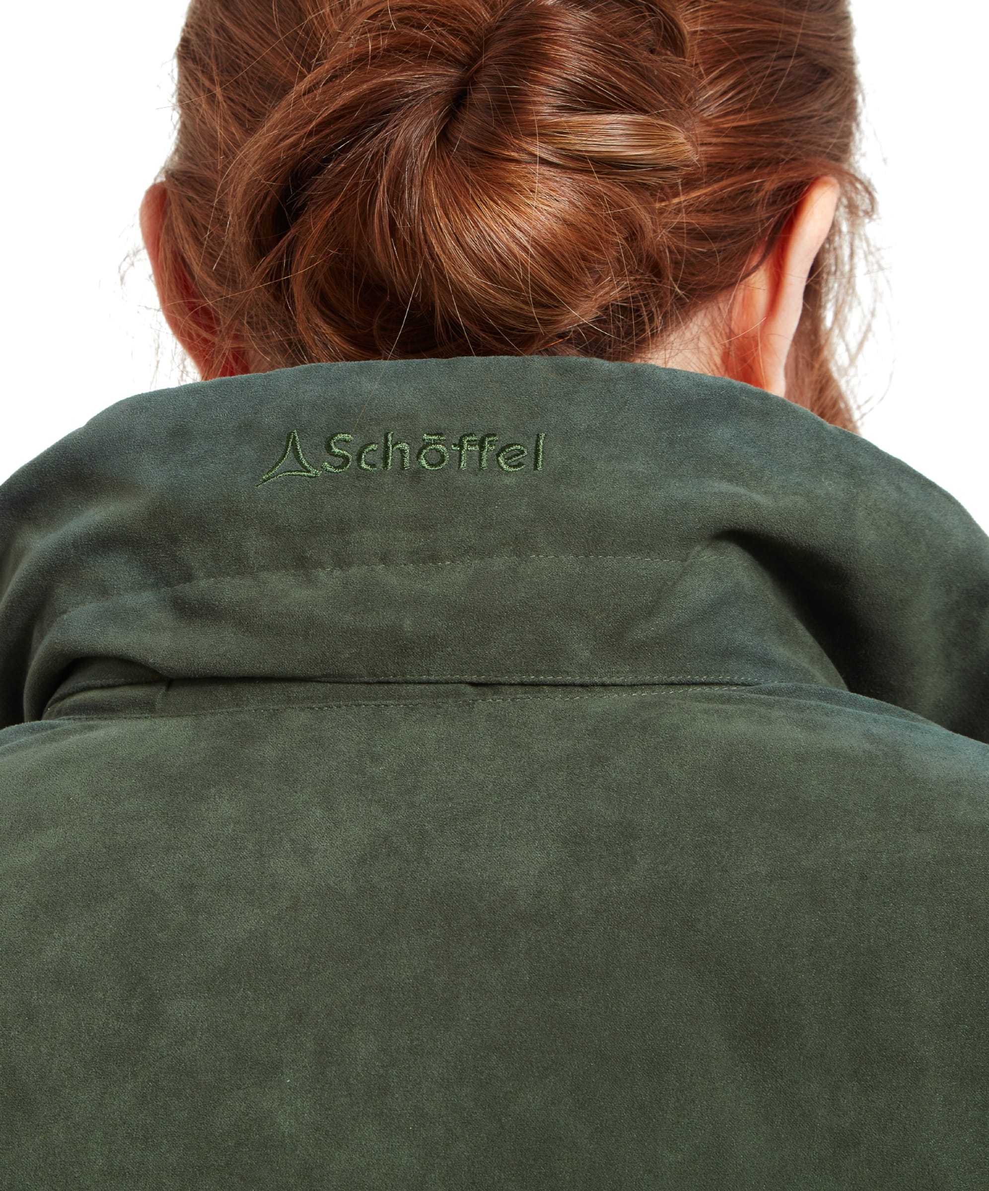 A close-up of the back of the Schöffel Teal Shooting Coat for Women in Green’s collar, showing the embroidered brand name “Schöffel” in matching green thread. The collar is raised, and the woman’s auburn hair is neatly tied up in a bun just above the collar.