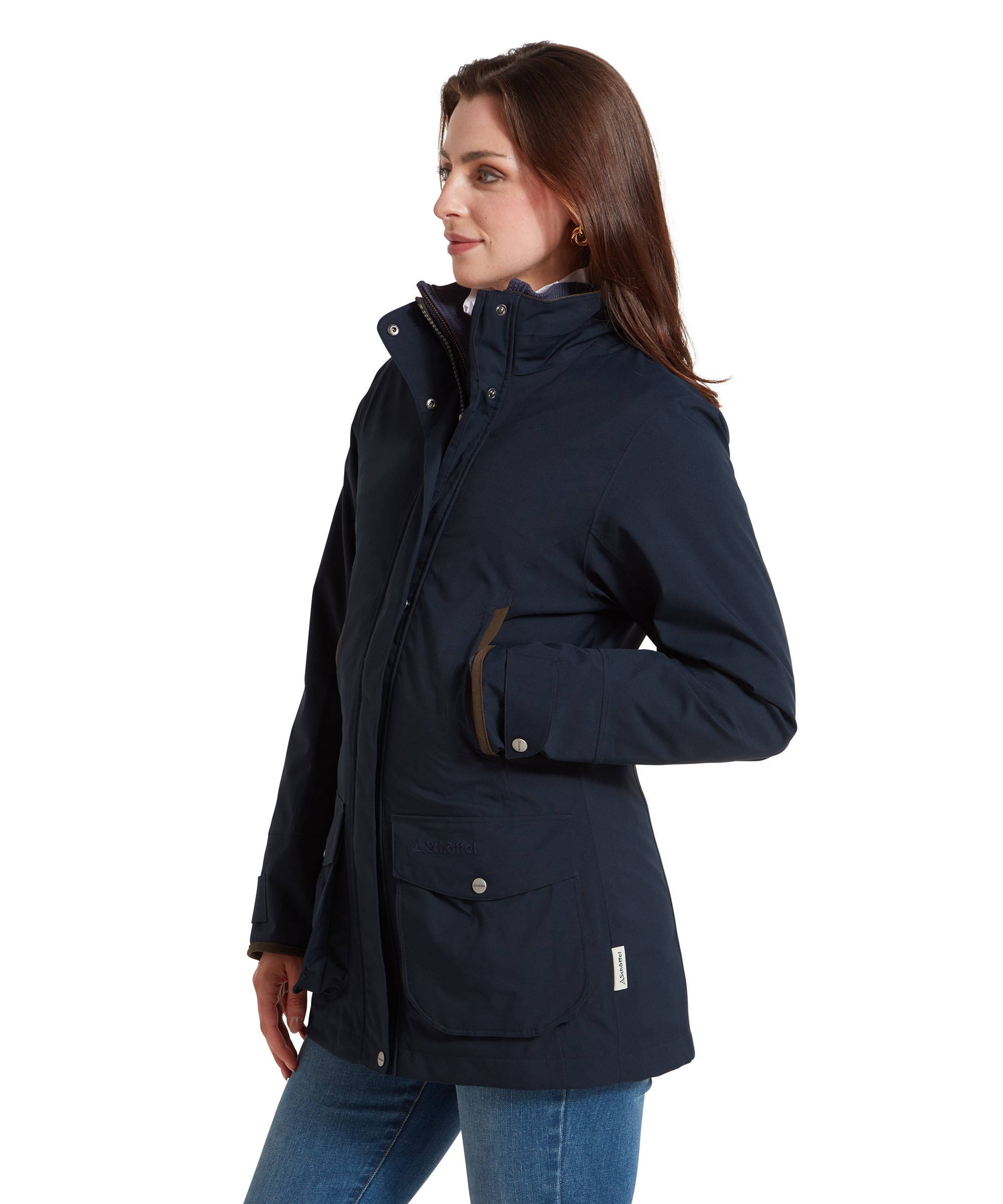 A side profile view of the woman in the Schöffel Teal Ultralight Coat for Women in Blue. She is facing left, with her hands in her jacket pockets. The jacket’s structured fit is evident, as well as the brown trim detail around the pockets and cuffs. The high collar is partially unzipped, showing the inner layer of her outfit.