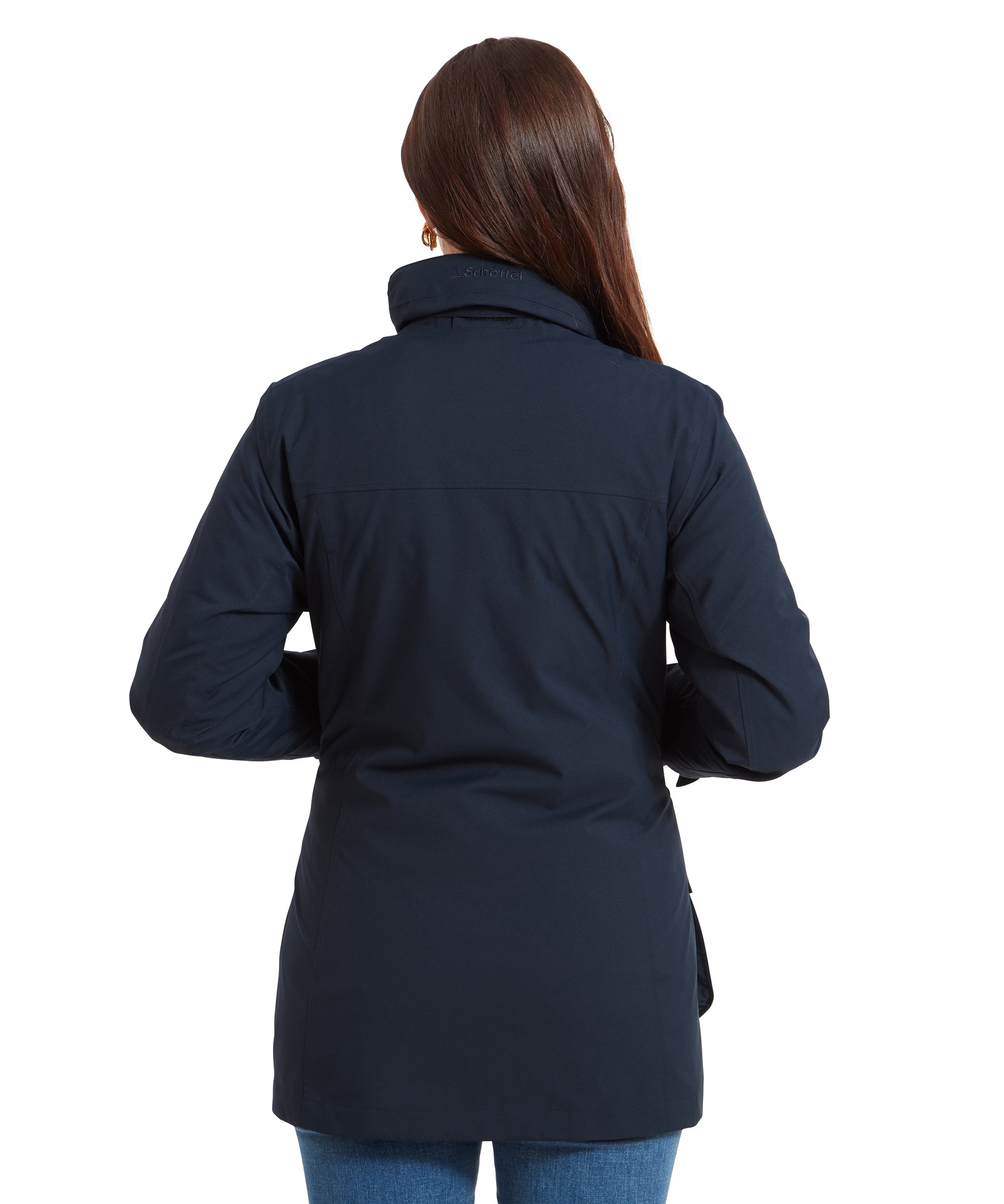 The woman is shown from the back, highlighting the rear design of the Schöffel Teal Ultralight Coat for Women in Blue. The jacket’s clean lines and tailored fit are visible, along with a subtle embroidered brand name, “Schöffel,” on the raised collar. Her straight brown hair falls loosely over her shoulders.