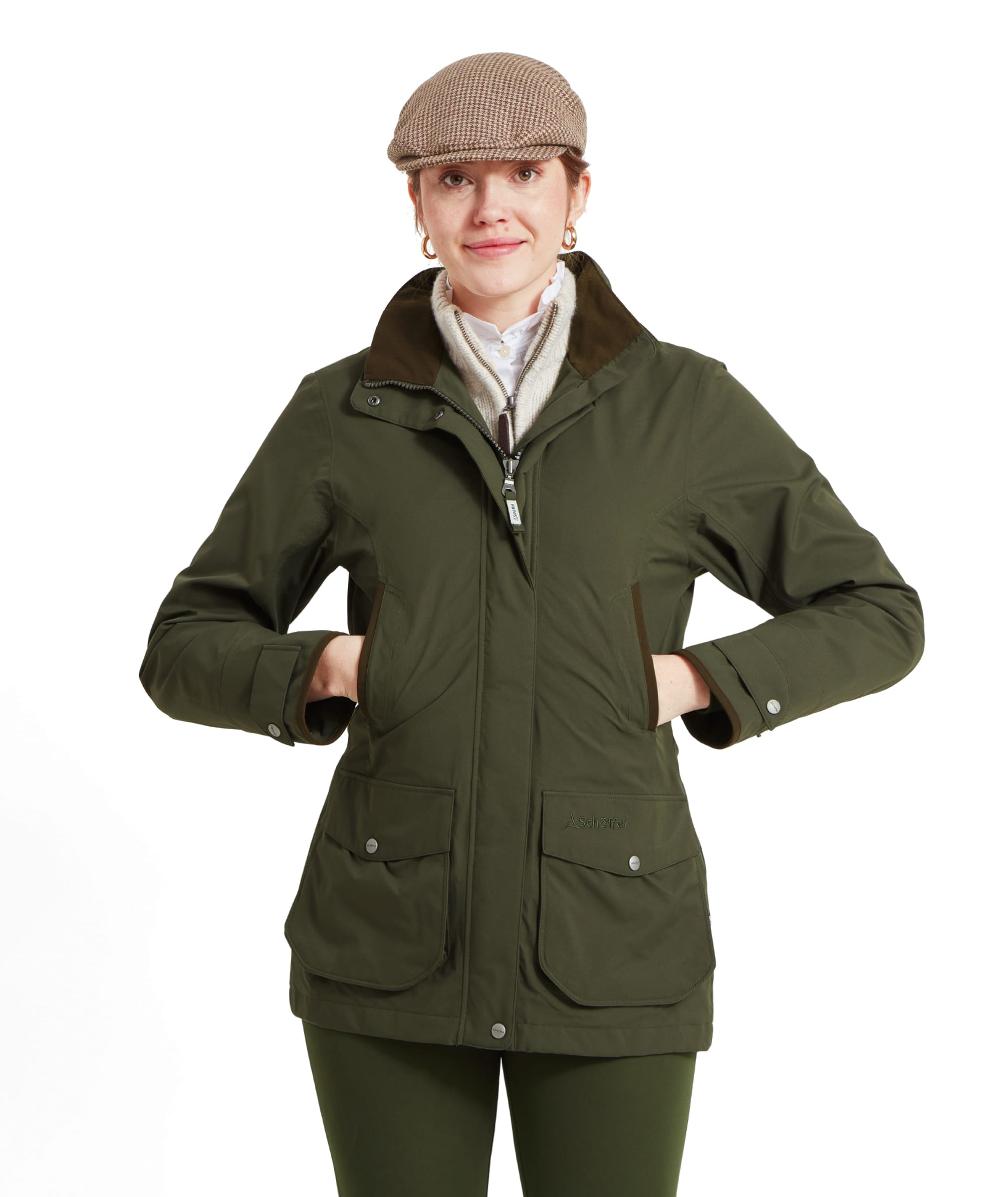 A woman is shown wearing a Schöffel Teal Ultralight Coat for Women in Green with a high collar and two large front pockets. She is also wearing a beige houndstooth flat cap, a white shirt, and a light grey zip-up sweater underneath the jacket. The woman is smiling slightly with her hands in the jacket's pockets, presenting a casual yet stylish look.