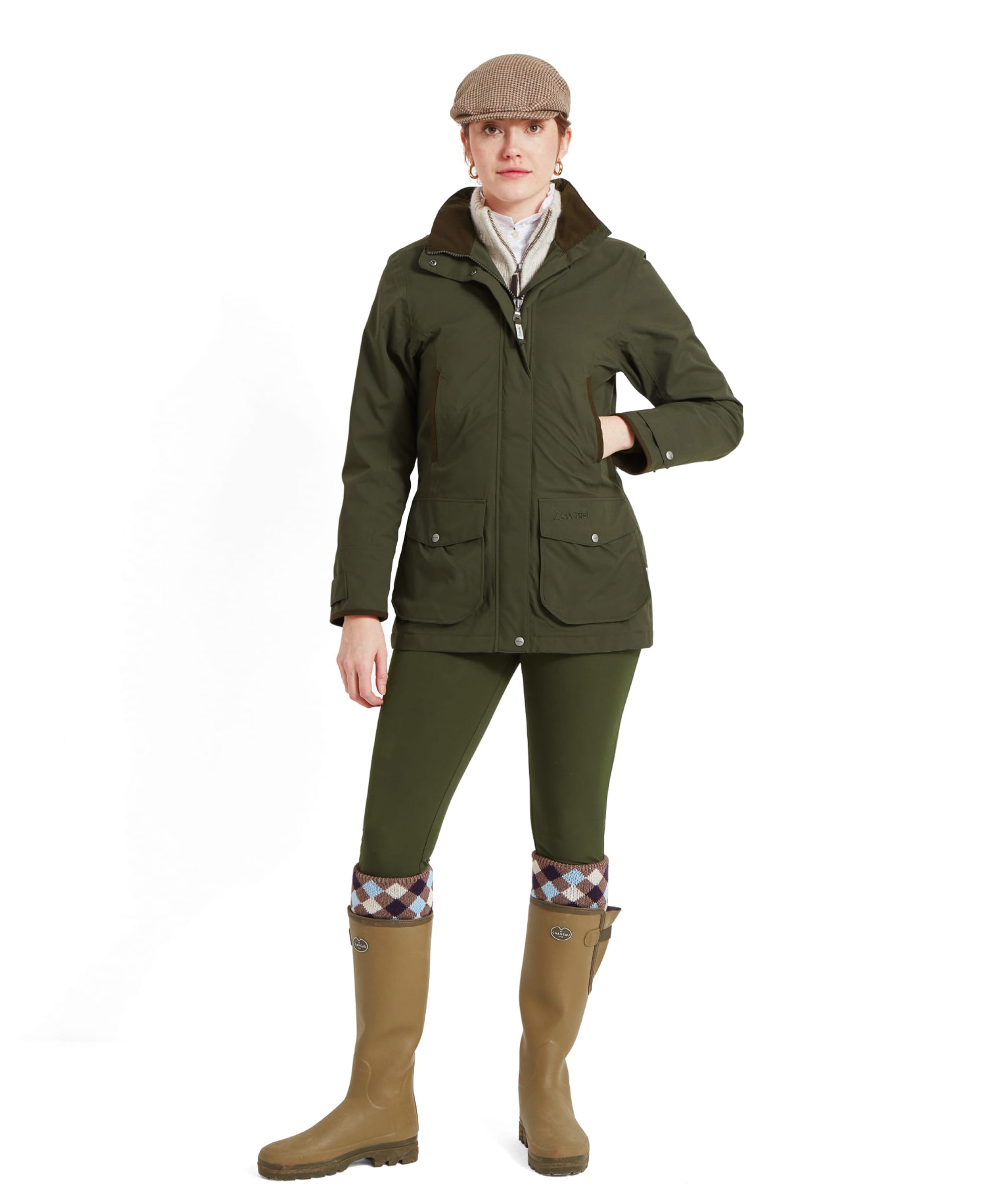 The same woman is depicted in a full-body shot wearing the Schöffel Teal Ultralight Coat for Women in Green, paired with matching green pants, brown wellington boots with patterned socks peeking out, and the beige houndstooth cap. She stands confidently with one hand in her pocket, showcasing the outdoor and functional style of the outfit.