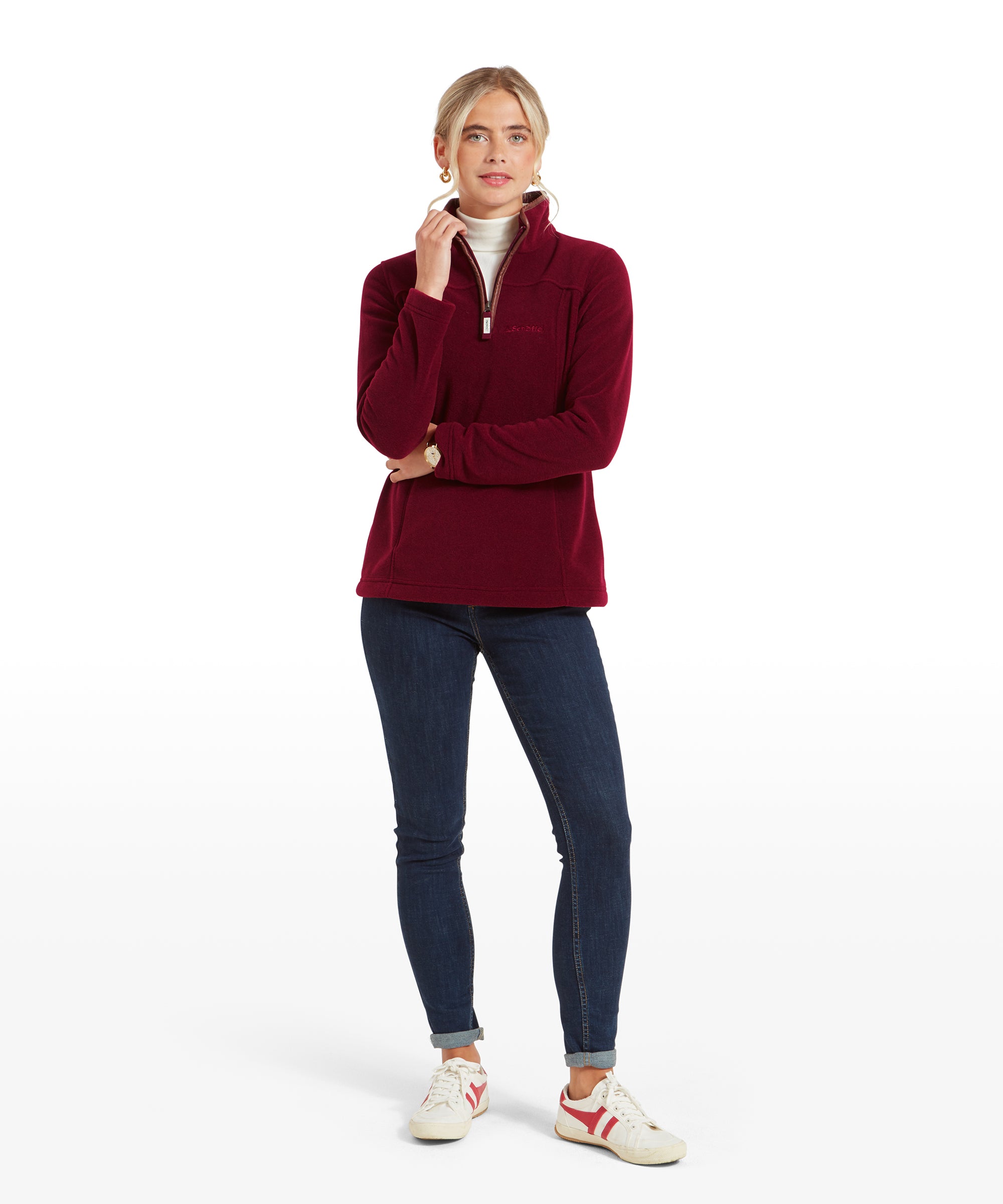 The same woman is depicted in a full-body shot, wearing the Schöffel Tilton 1/4 Zip Fleece for Women in Red, dark blue skinny jeans, and white sneakers with red accents. She stands with one hand gently touching her chin and the other at her side, creating a thoughtful and relaxed pose. The white turtleneck peeks out from under the fleece, adding a layer of warmth to the outfit.