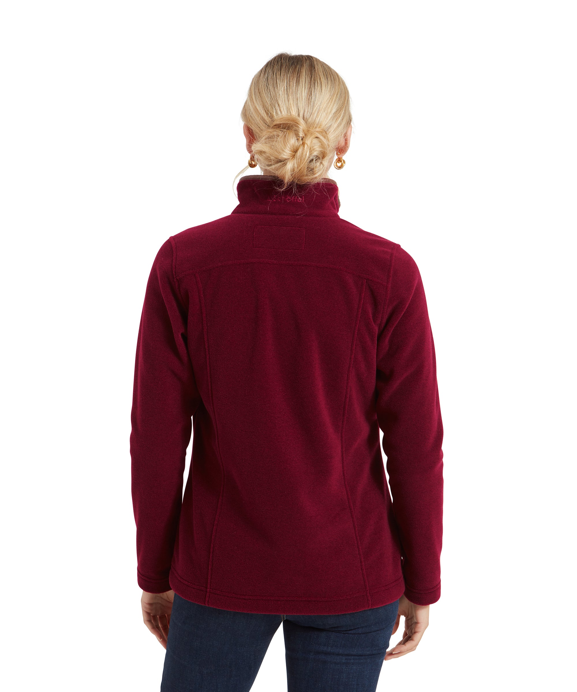 The woman is shown from behind, highlighting the clean and simple design of the red fleece. The back has subtle seam details, and the collar stands upright. Her blonde hair is styled in a neat, low bun, secured with a black hair tie, and she wears gold earrings that add a bit of sparkle.