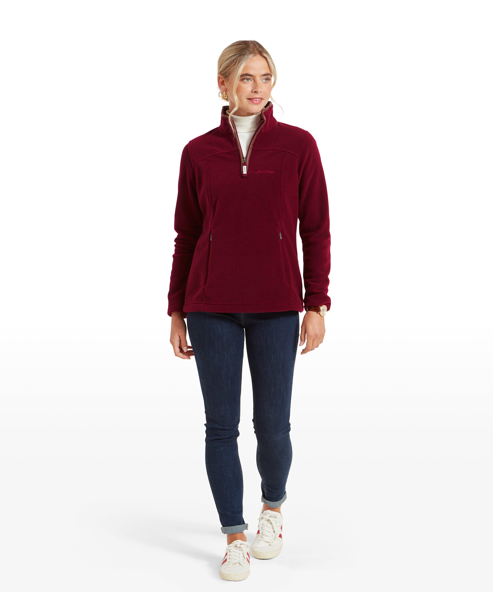 In this image, the woman is walking forward, wearing the red fleece and dark blue skinny jeans. She pairs the outfit with white trainers that have red accents, which complement the fleece. She gazes off to the side with a slight smile, conveying a sense of ease and confidence.