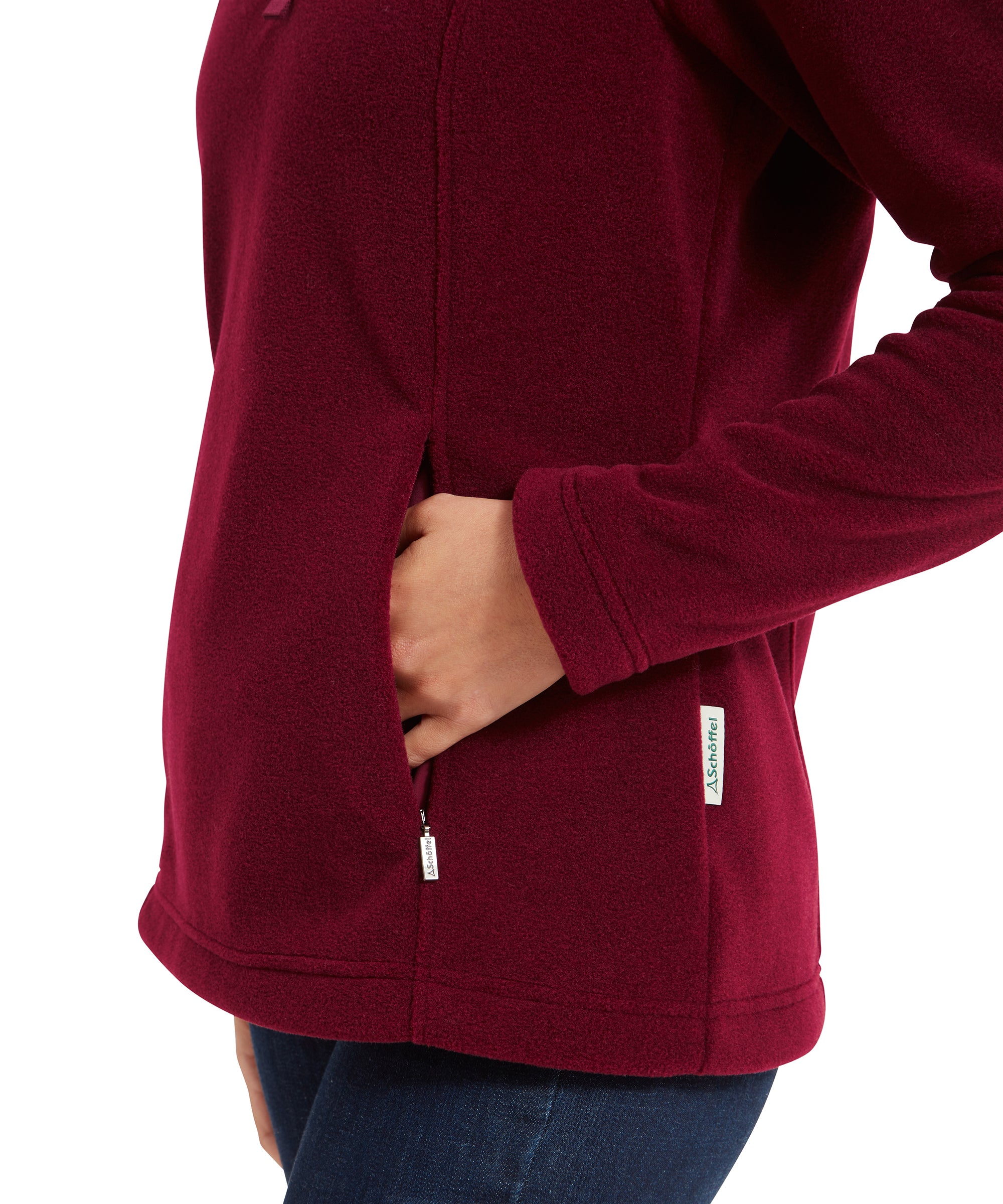 A close-up shot focuses on the woman’s hand tucked into one of the fleece’s side pockets. The deep red fabric is contrasted by her dark blue jeans, and a small brand tag is visible near the pocket. The woman’s casual pose highlights the fleece’s practicality and comfort.