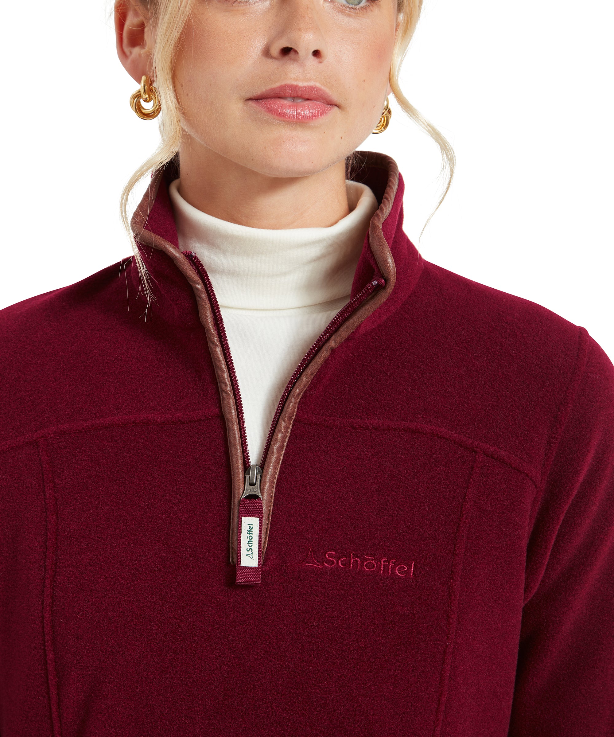 This image zooms in on the front of the fleece, showing the brown trim around the collar and zipper. The high collar is slightly open, revealing the white turtleneck underneath. The brand’s logo is embroidered on the chest, and the zipper pull is labeled with the brand name, showcasing the attention to detail in the design.